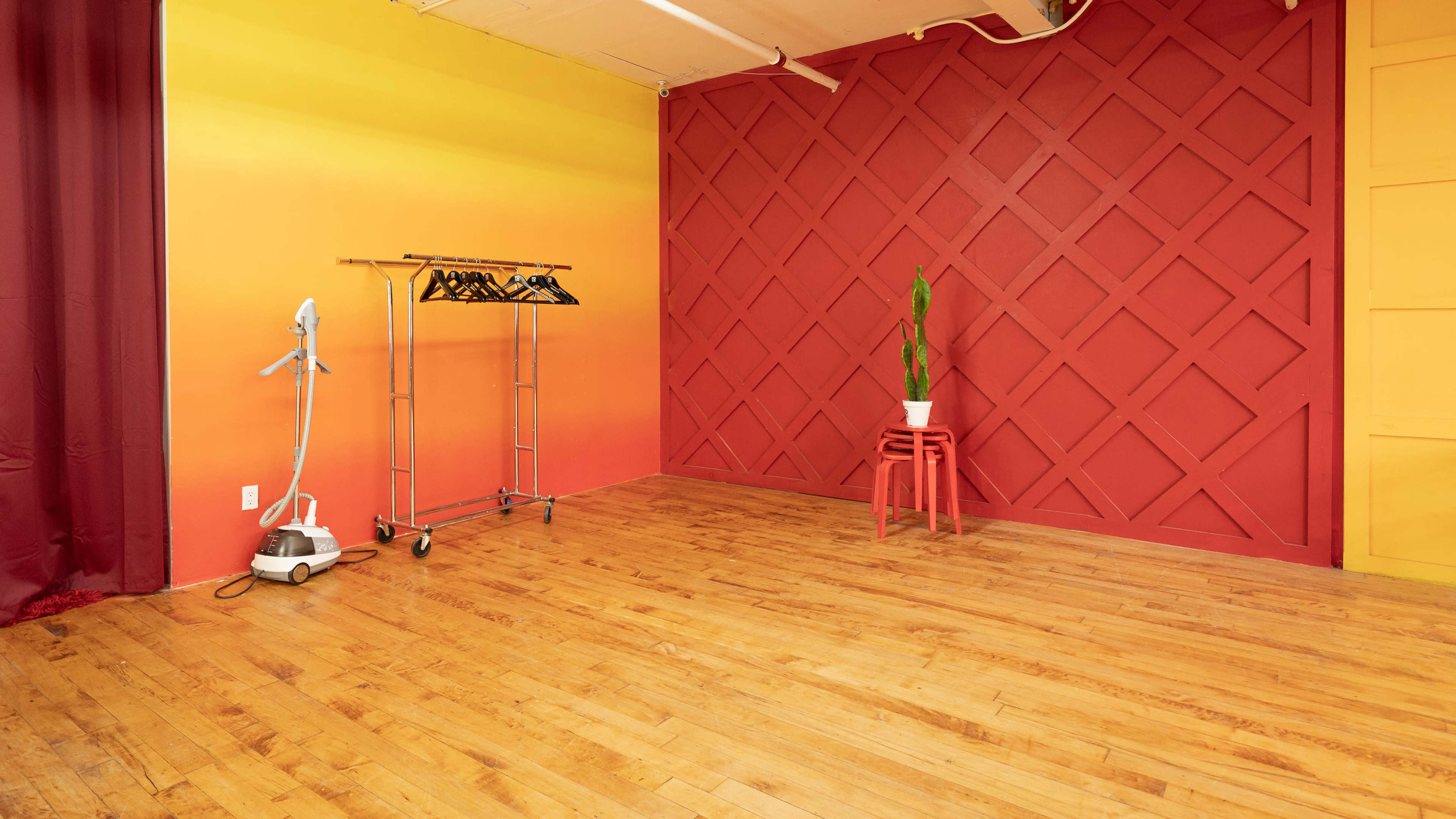 A room with a gradient yellow to orange wall, a red accent wall with a diamond pattern, a clothing rack, and a potted plant on a small red stool.