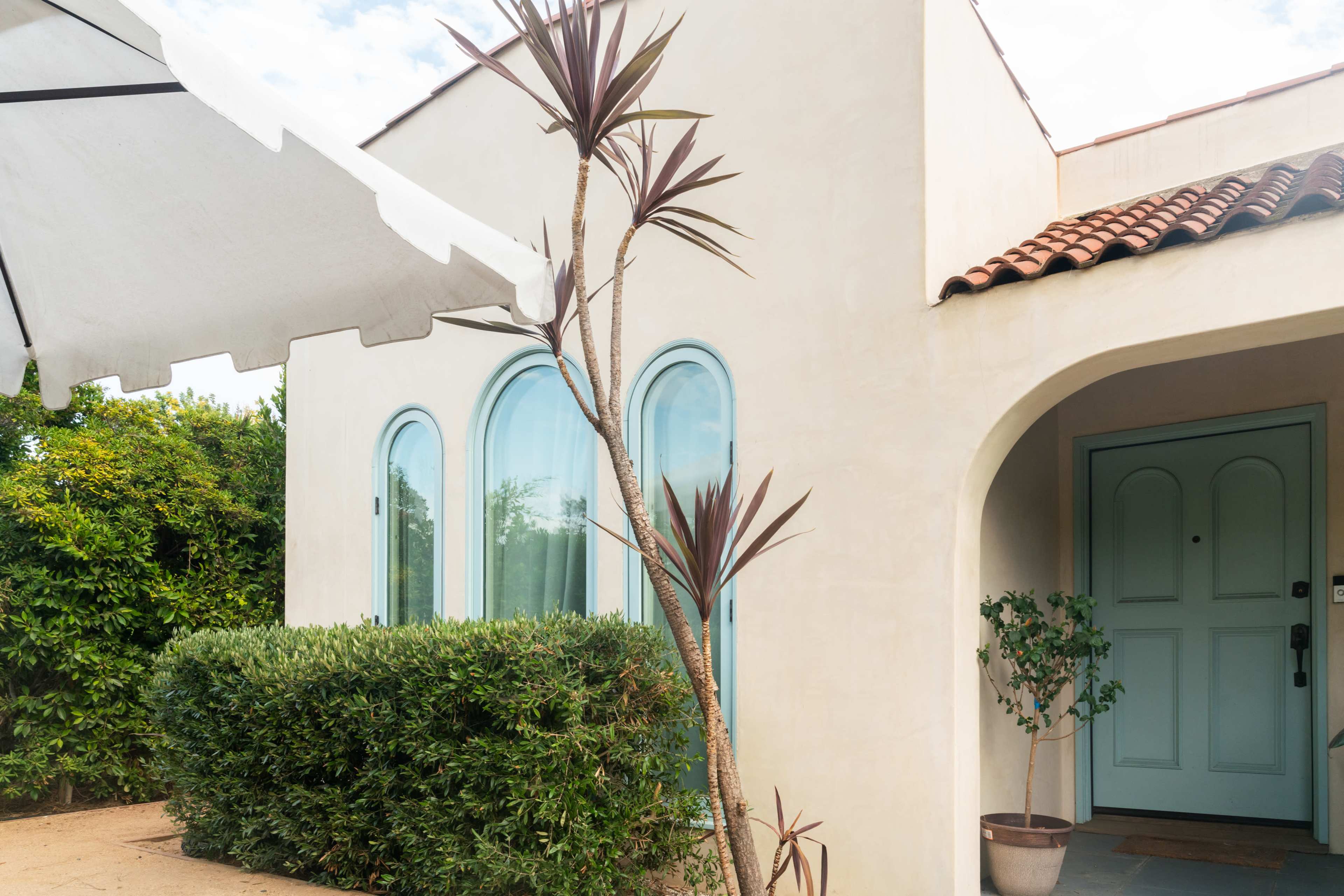 A light-colored stucco house features arched windows, a green door, and a potted plant near the entrance, with landscaping visible in the background.