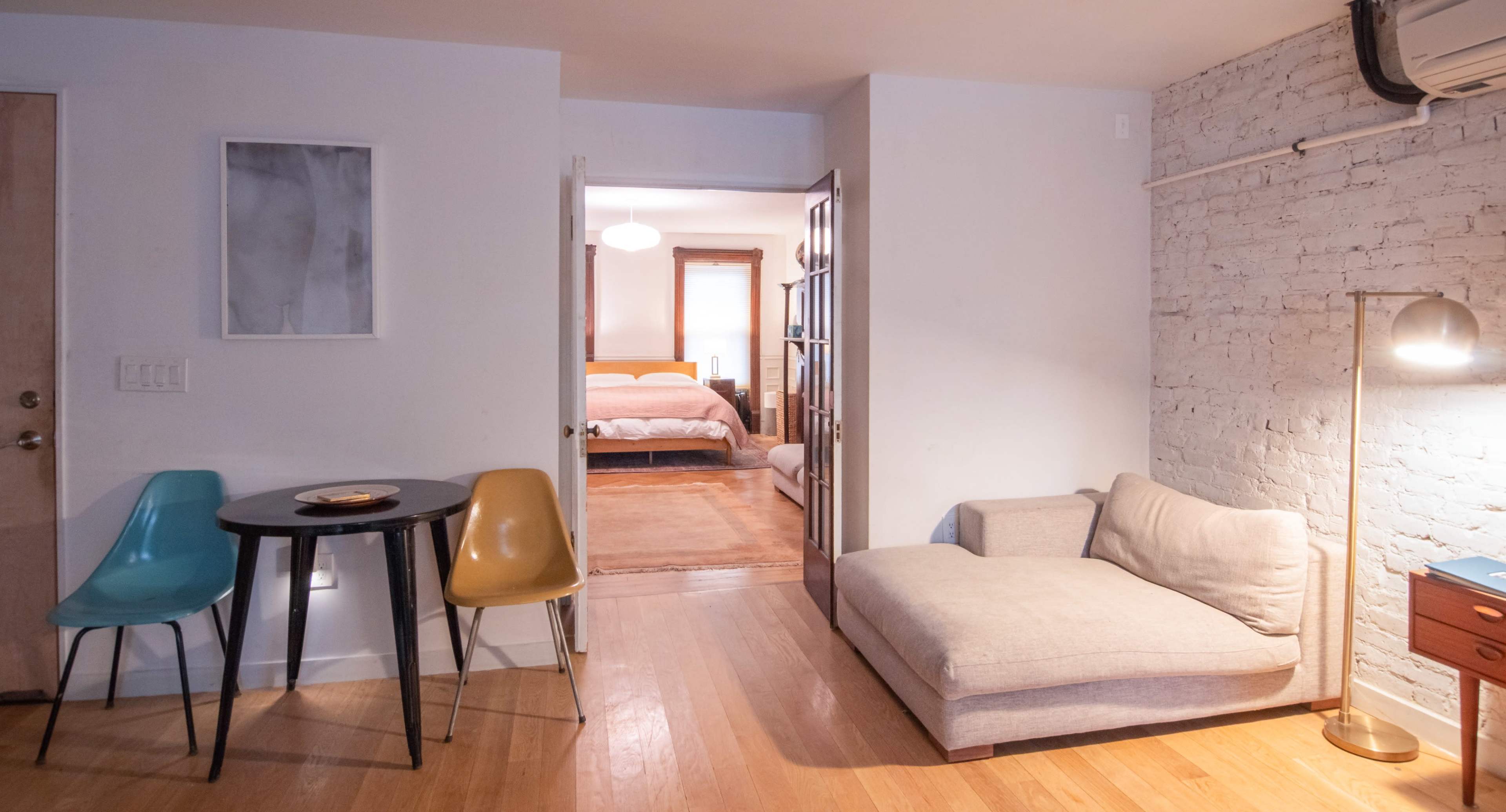 The image shows a small living space featuring a table with two chairs, a couch, and an open doorway leading to a bedroom.