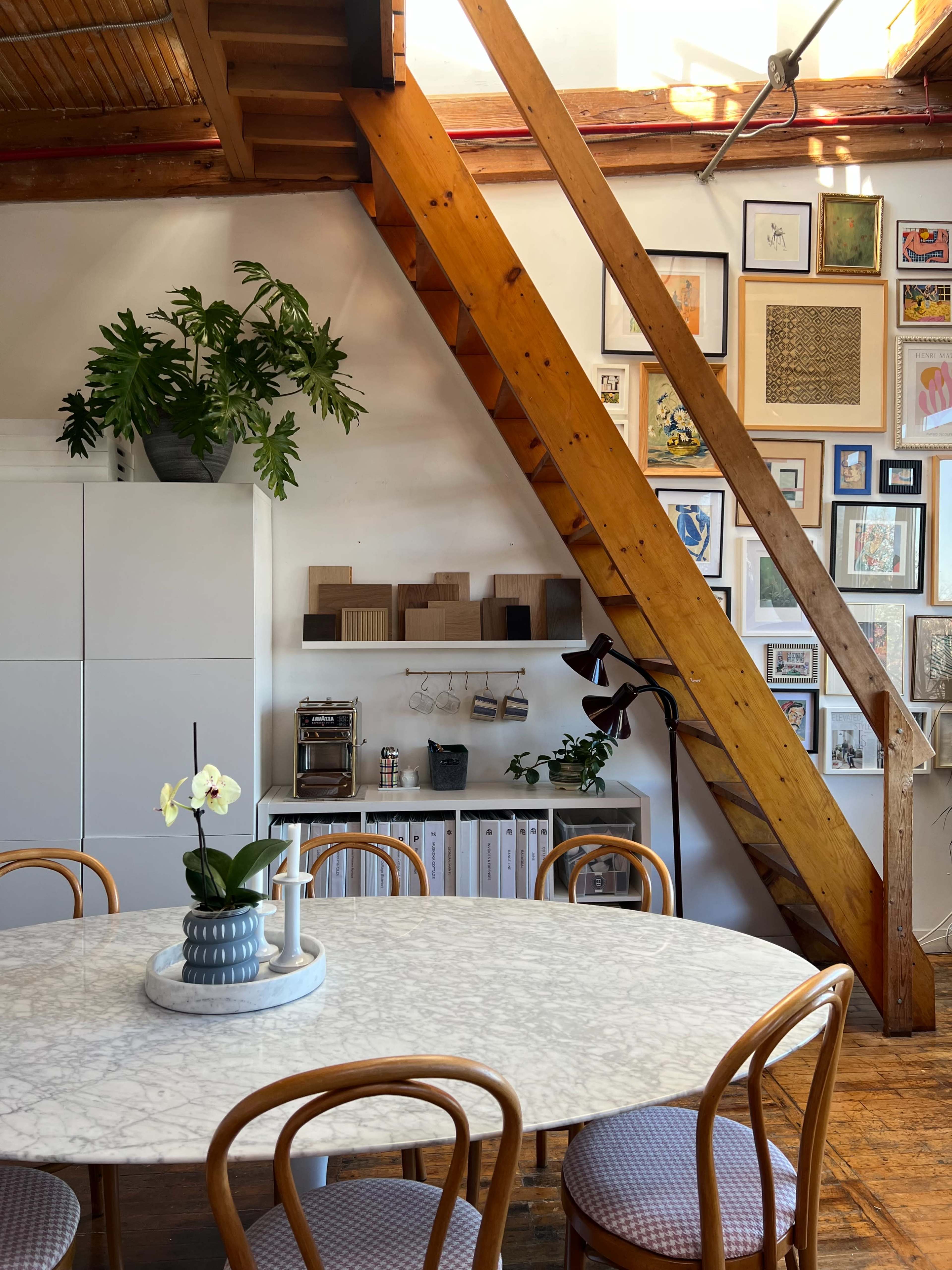 A dining area features a round marble table surrounded by bentwood chairs, with a wooden beam structure and a wall adorned with various framed artworks.
