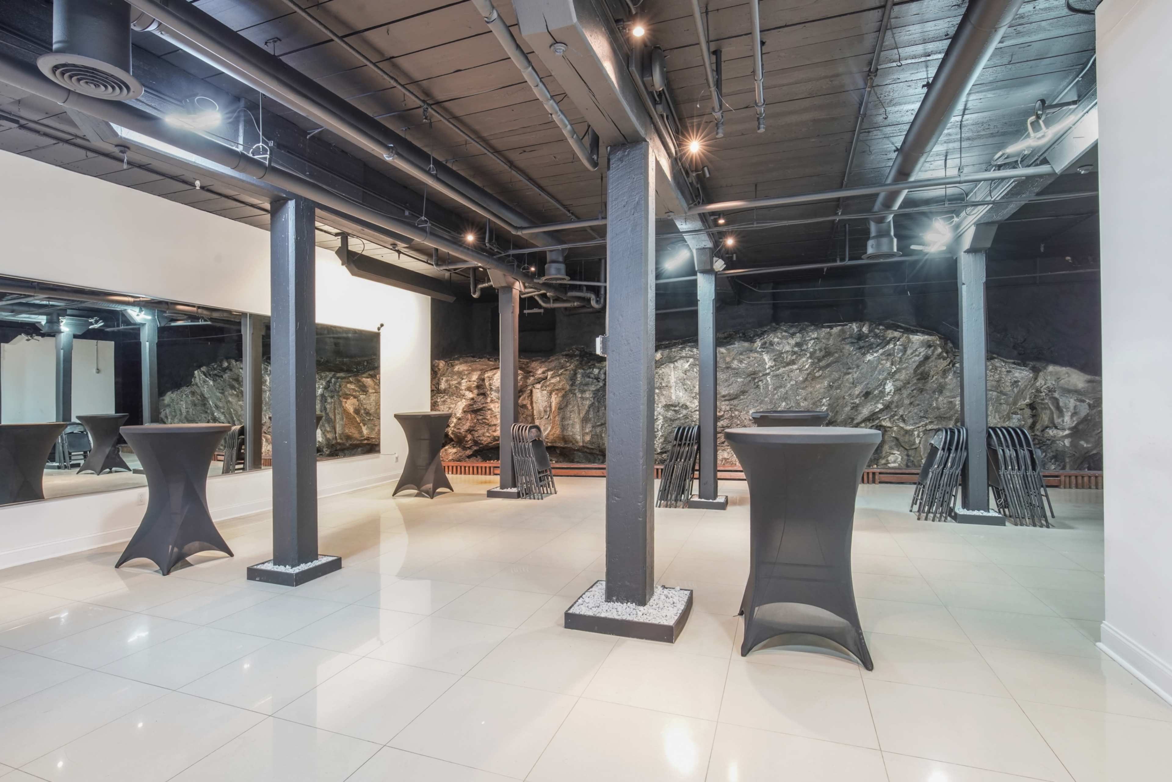 The image shows a spacious venue with dark columns and sleek tables, featuring a rock wall in the background.