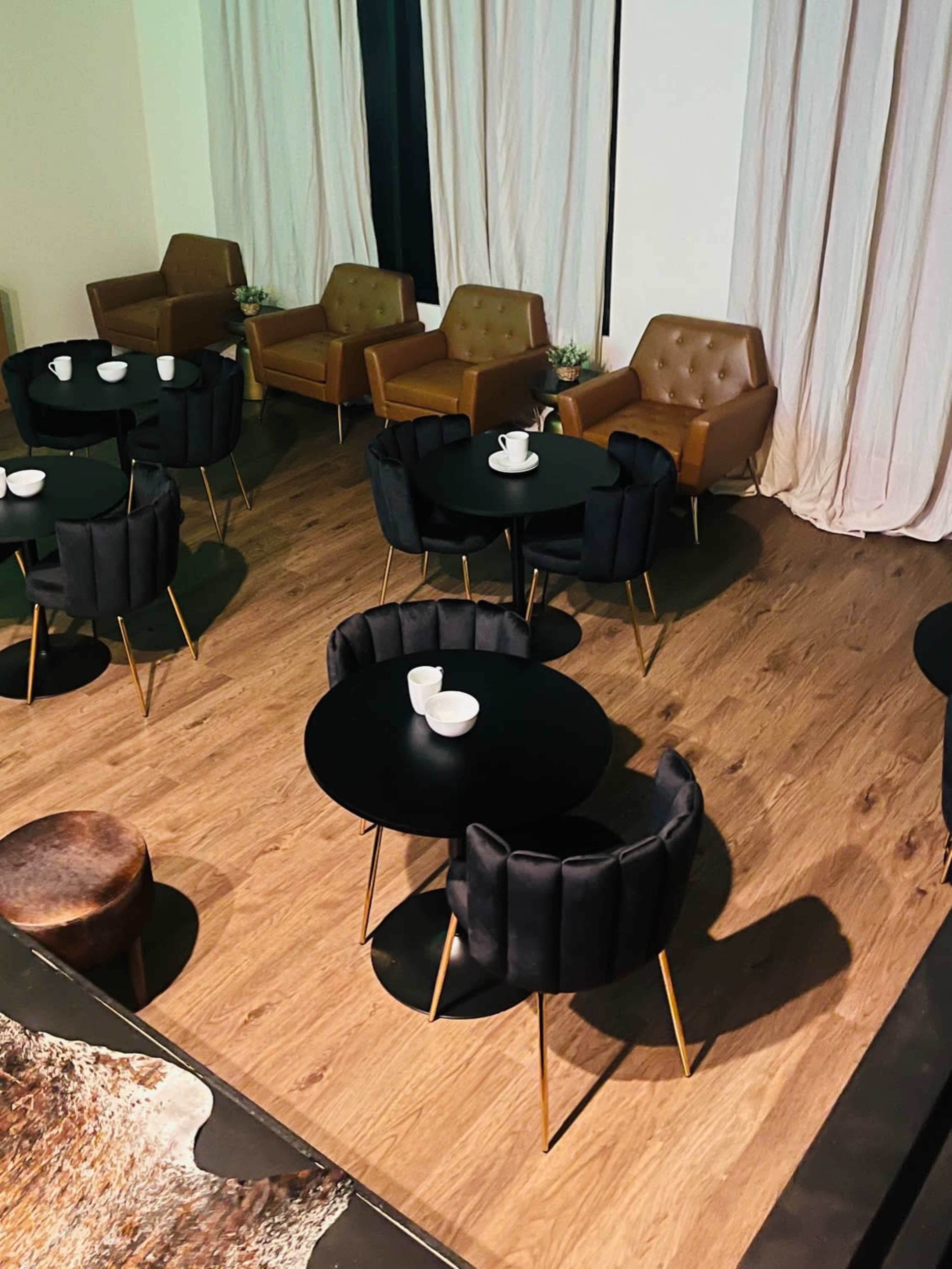 A modern café interior with wooden flooring, several round black tables surrounded by upholstered chairs, and light-colored curtains in the background.