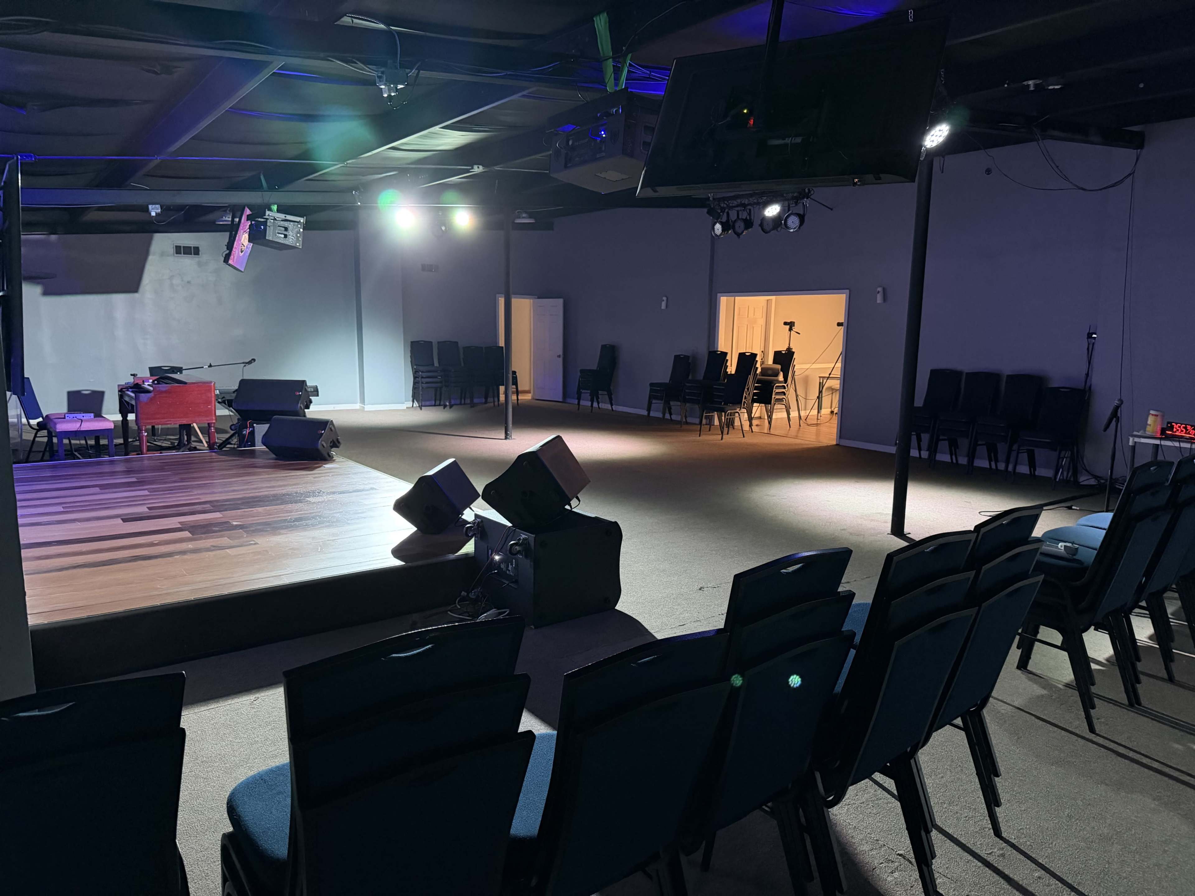 The image shows an empty auditorium with a stage at the front, surrounded by rows of blue chairs and dim lighting.