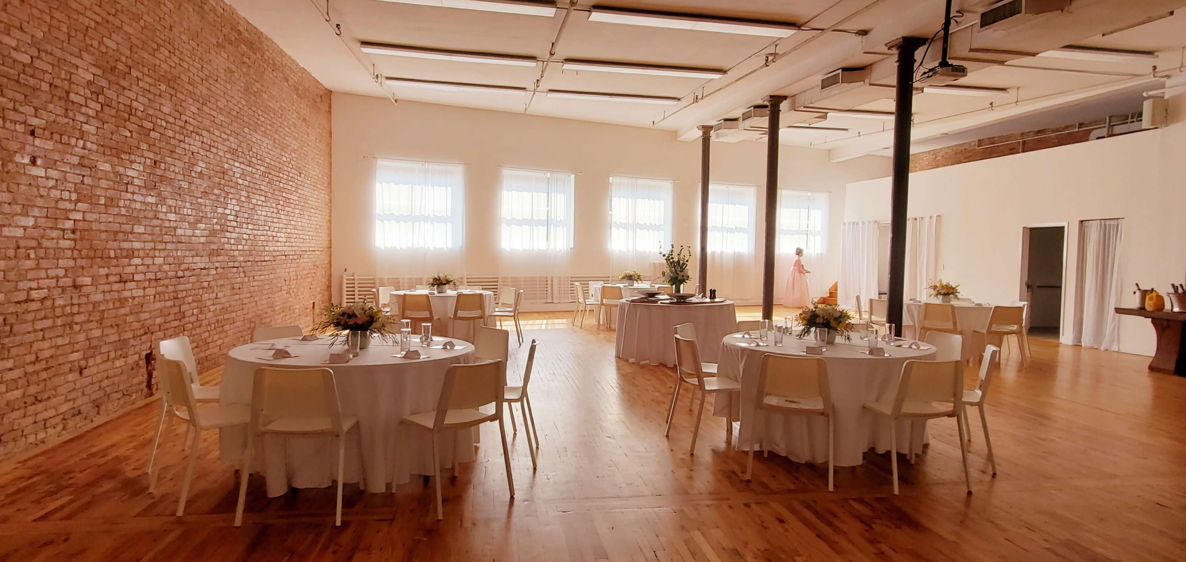 The image shows a spacious event venue with round tables set for dining, white tablecloths, and large windows illuminating the brick and wooden interior.