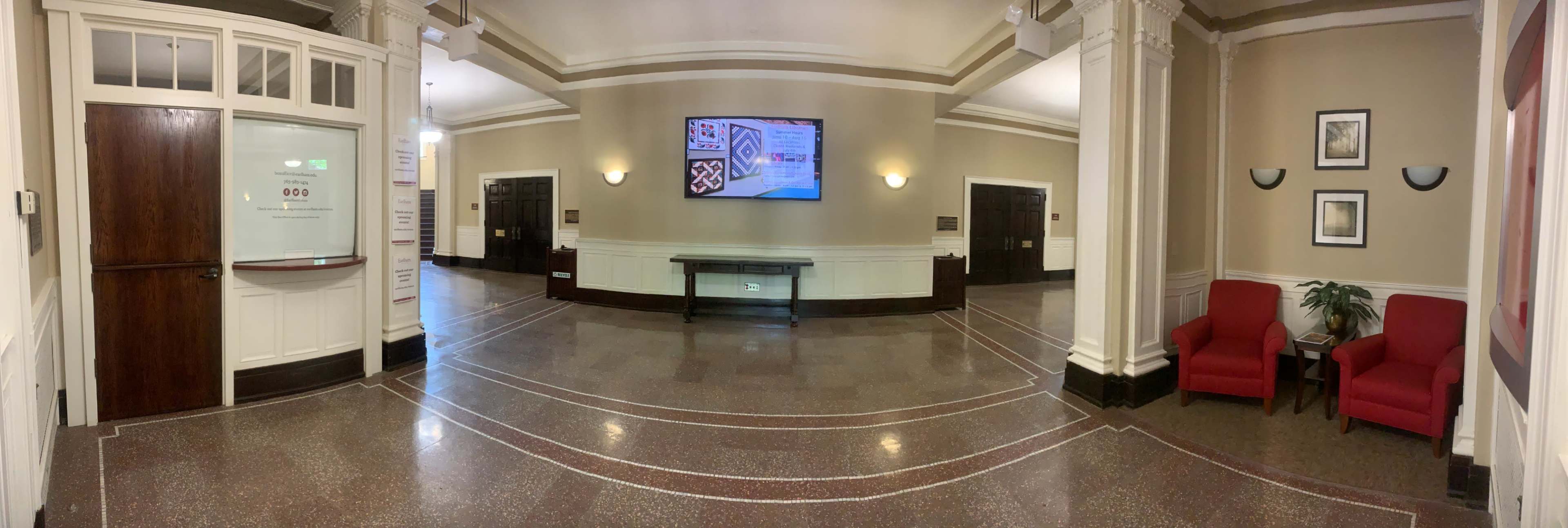 The image displays a spacious lobby area featuring a large screen on the wall, a reception counter to the left, and two red chairs positioned against the right wall.