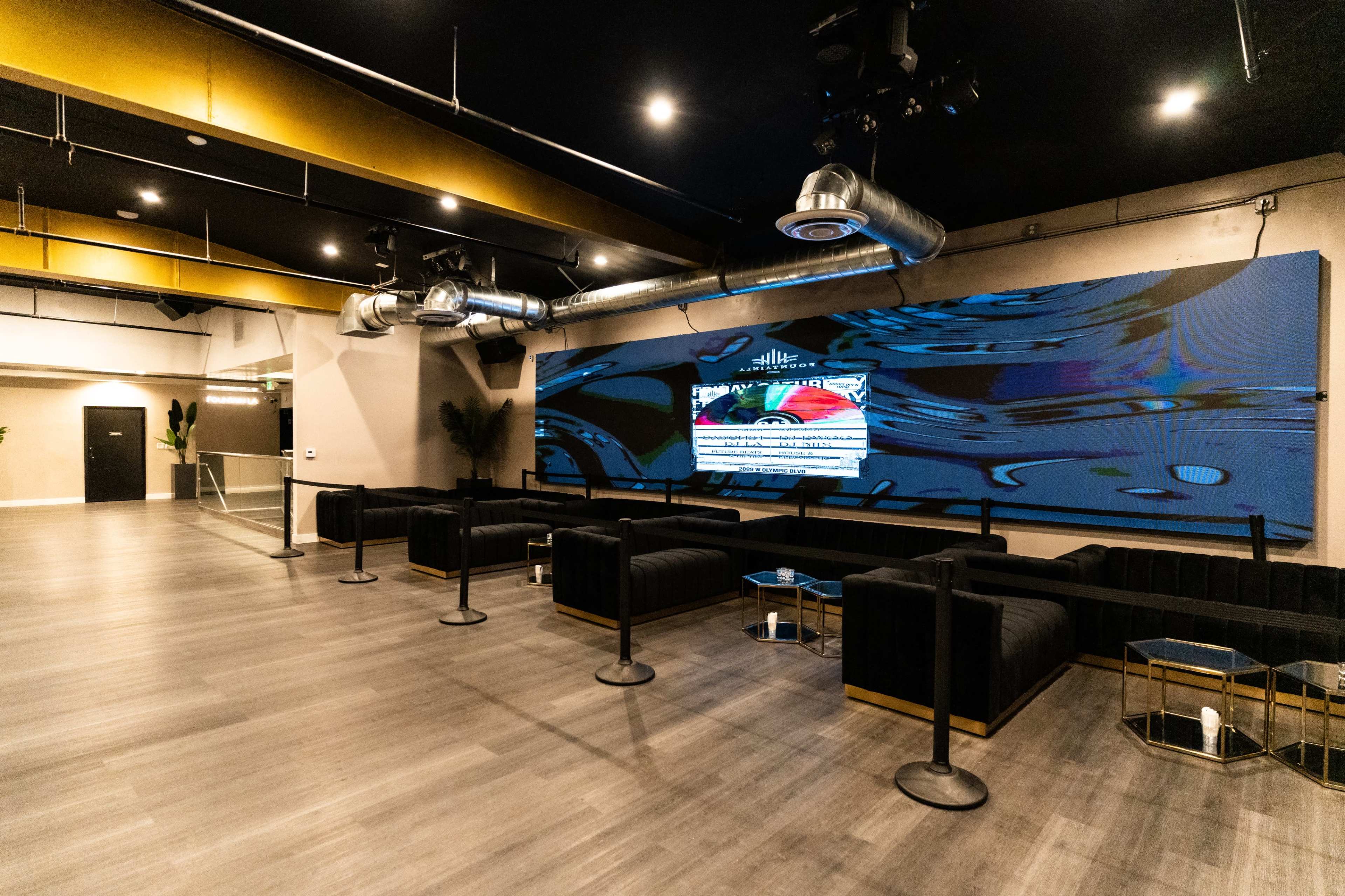 The image shows a modern lounge area featuring black seating, a large screen displaying visuals on a wall, and an open layout with a wooden floor.