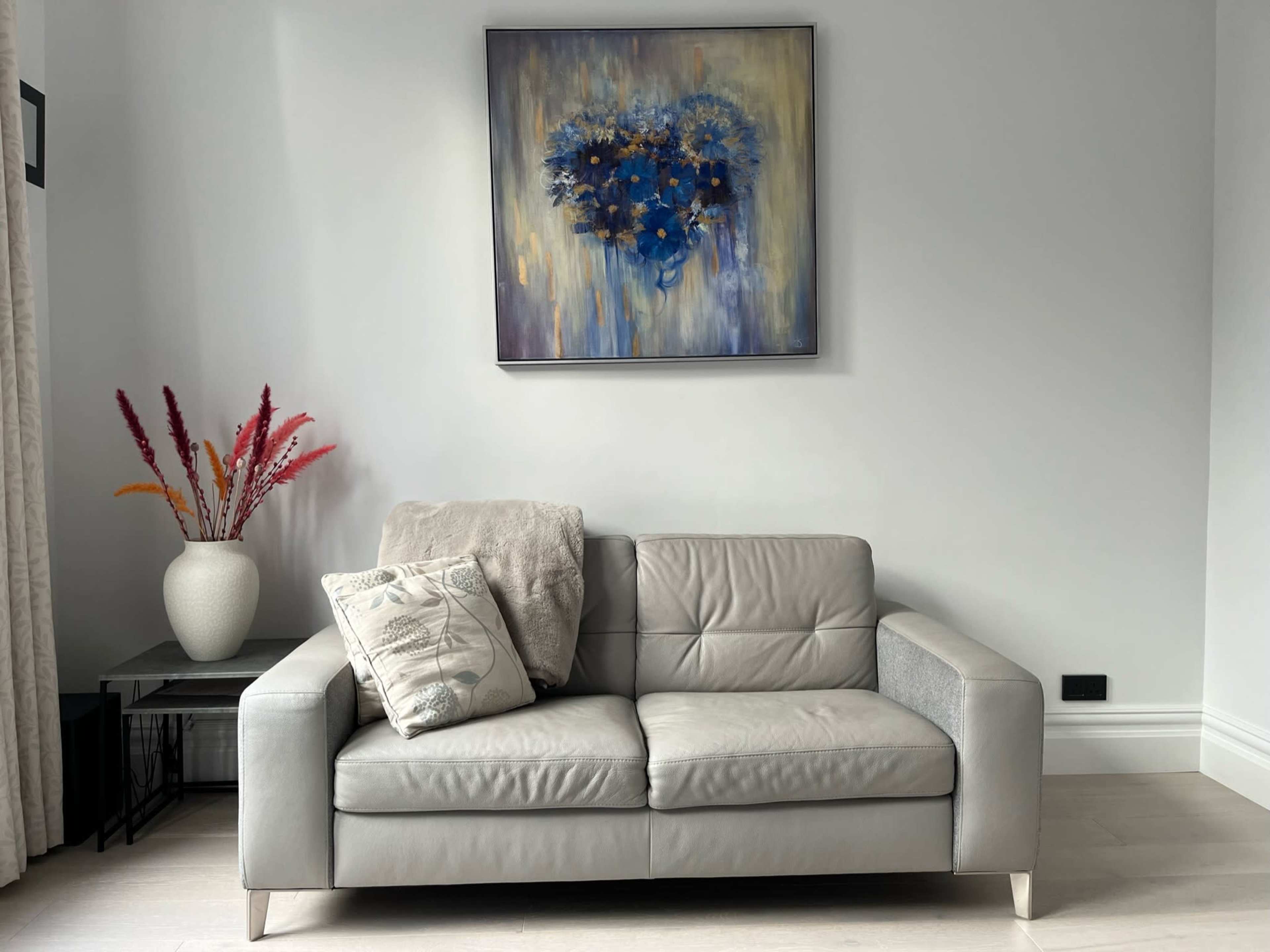 A light gray sofa with a decorative pillow sits against a pale wall, beneath a framed blue floral painting, alongside a vase of colorful dried flowers.