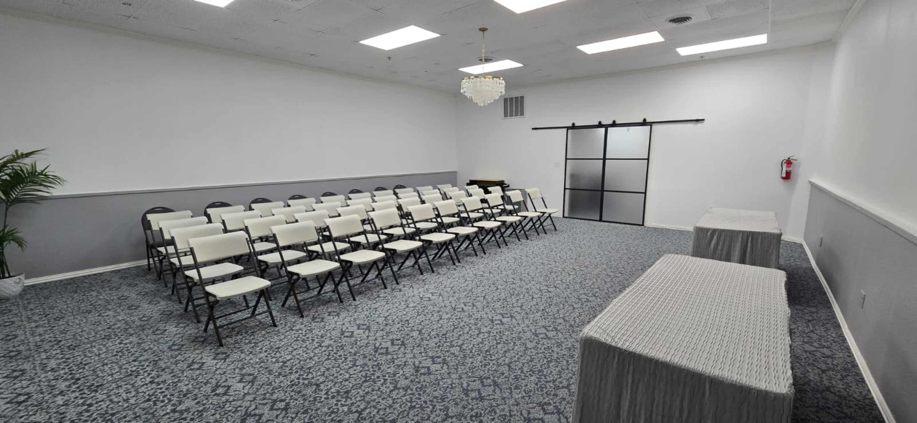 The image shows a room set up with rows of white chairs facing a wall, with two covered tables at one end and a glass door on the opposite side.