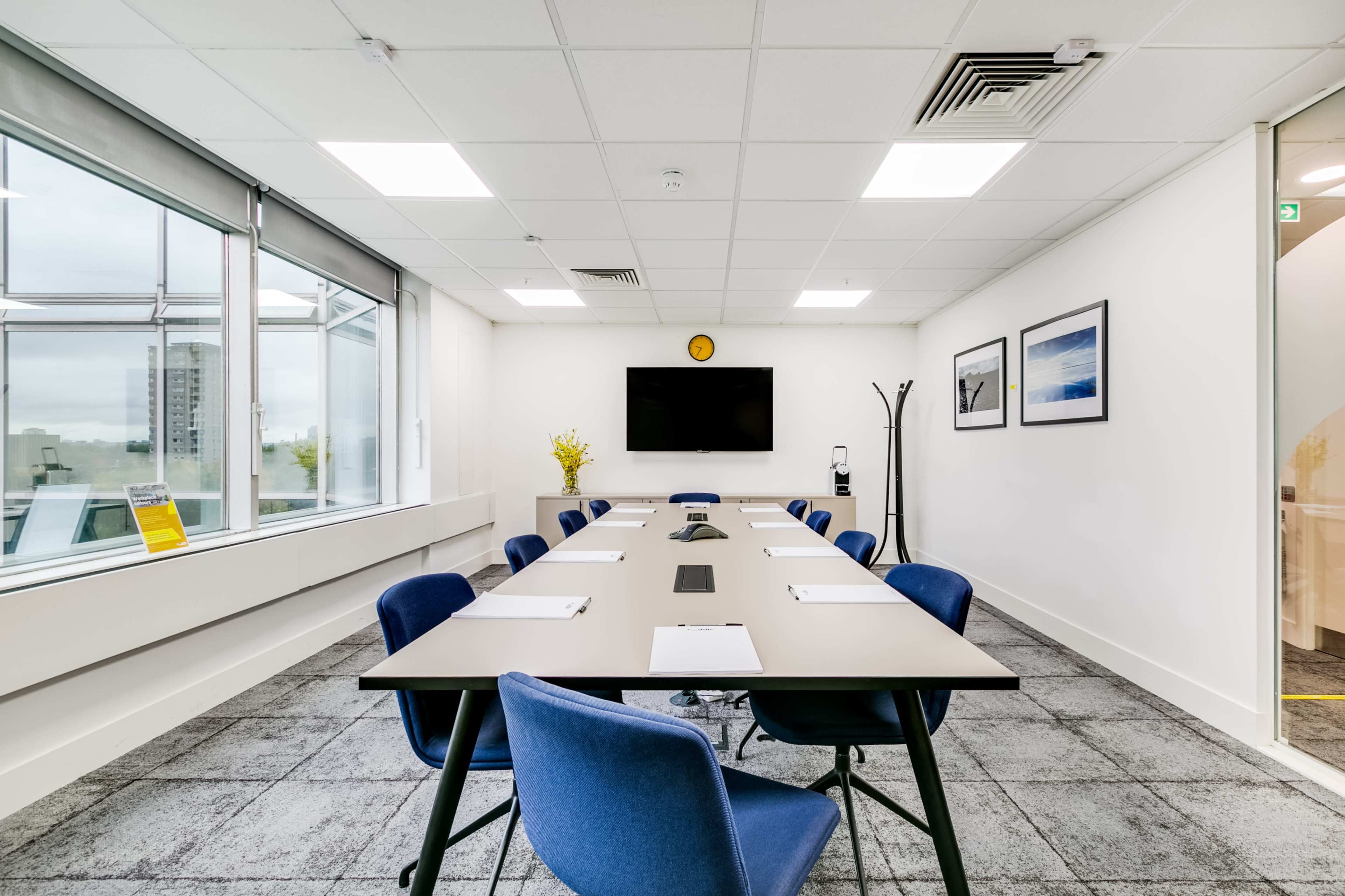 A modern conference room features a large table with blue chairs, a wall-mounted TV, and windows providing natural light.