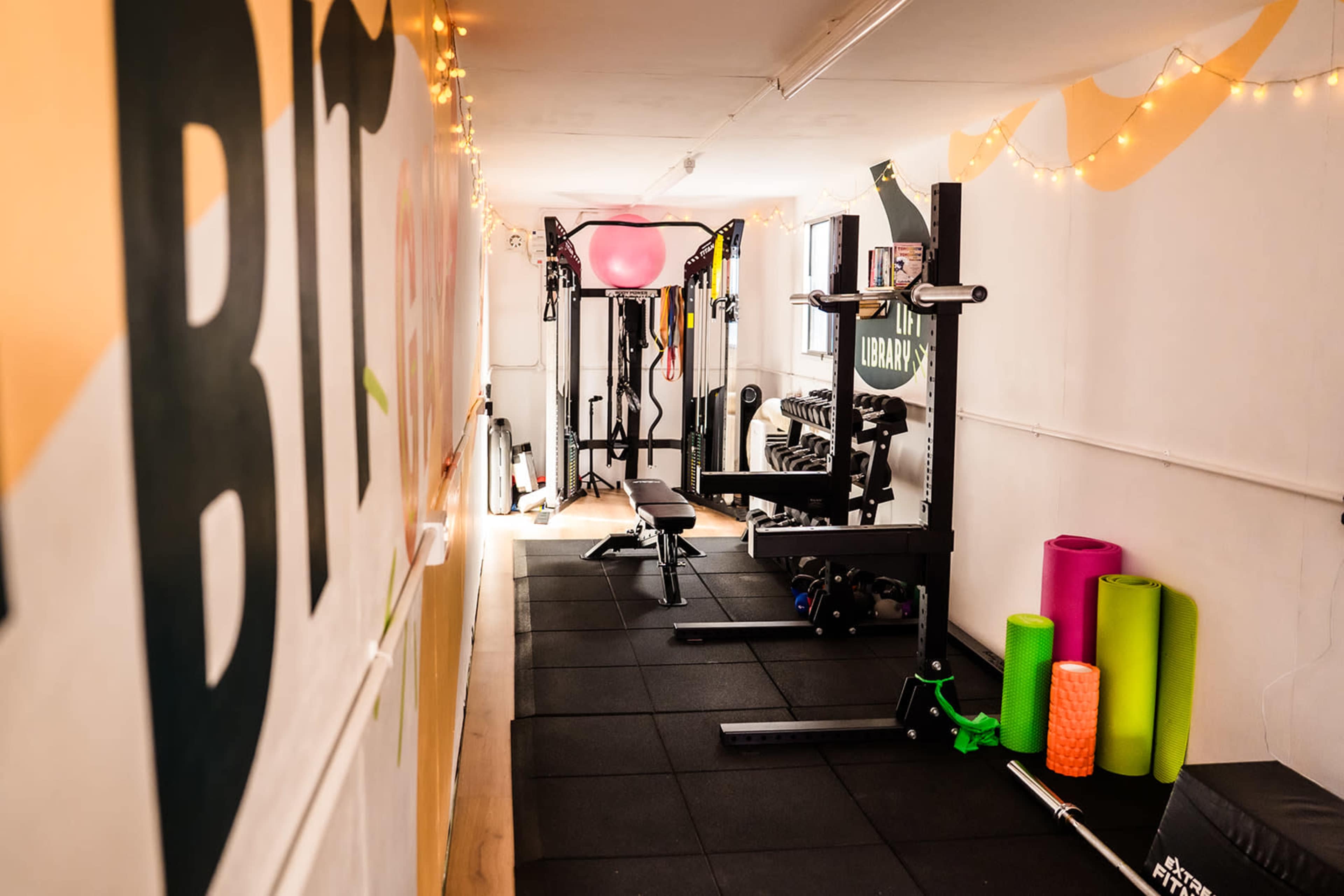 The image shows a narrow gym space with various exercise equipment, including a squat rack, weights, and fitness mats, illuminated by string lights along the wall.