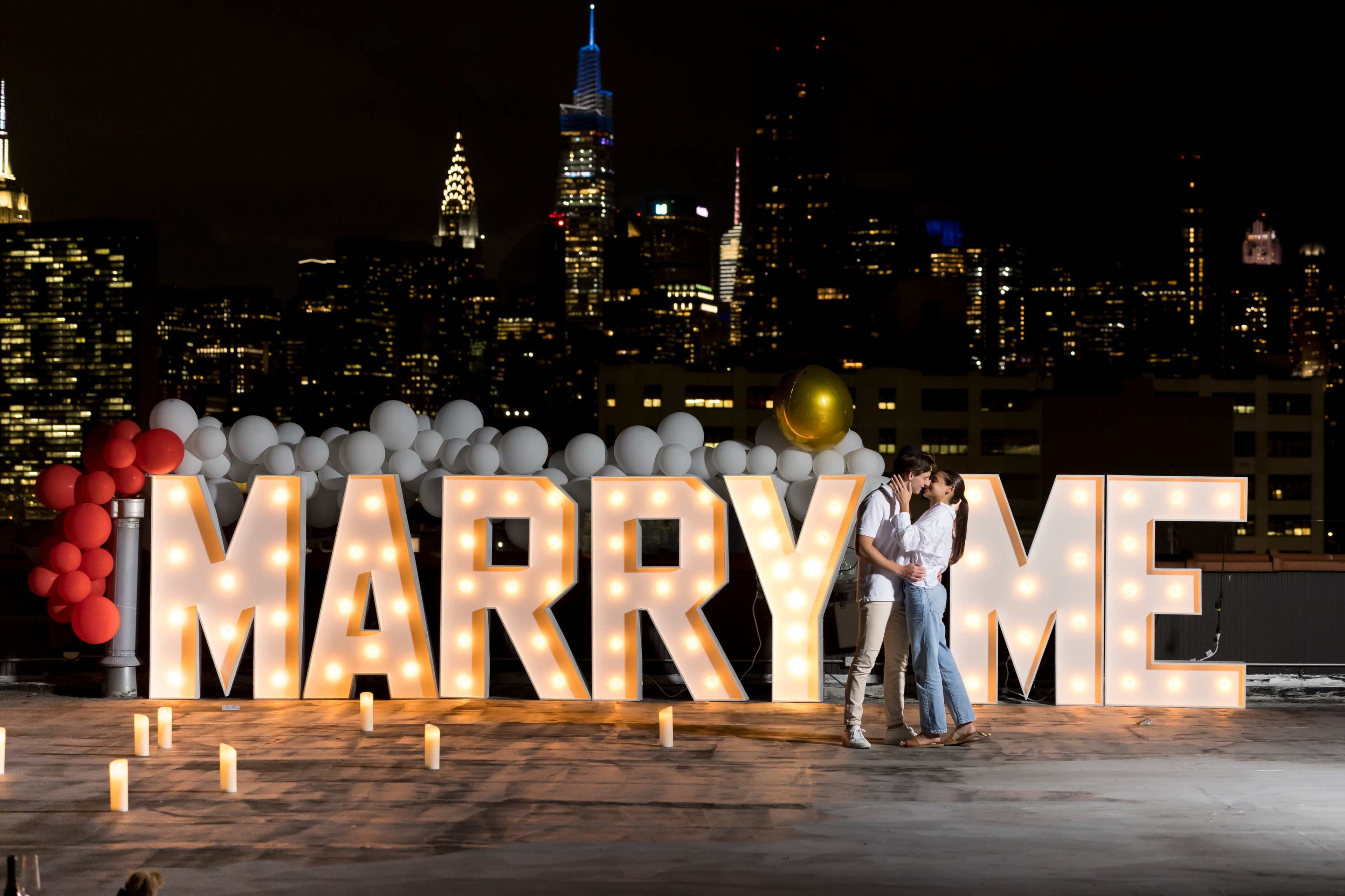 A couple stands on a rooftop illuminated by a large "MARRY ME" display against a city skyline at night.