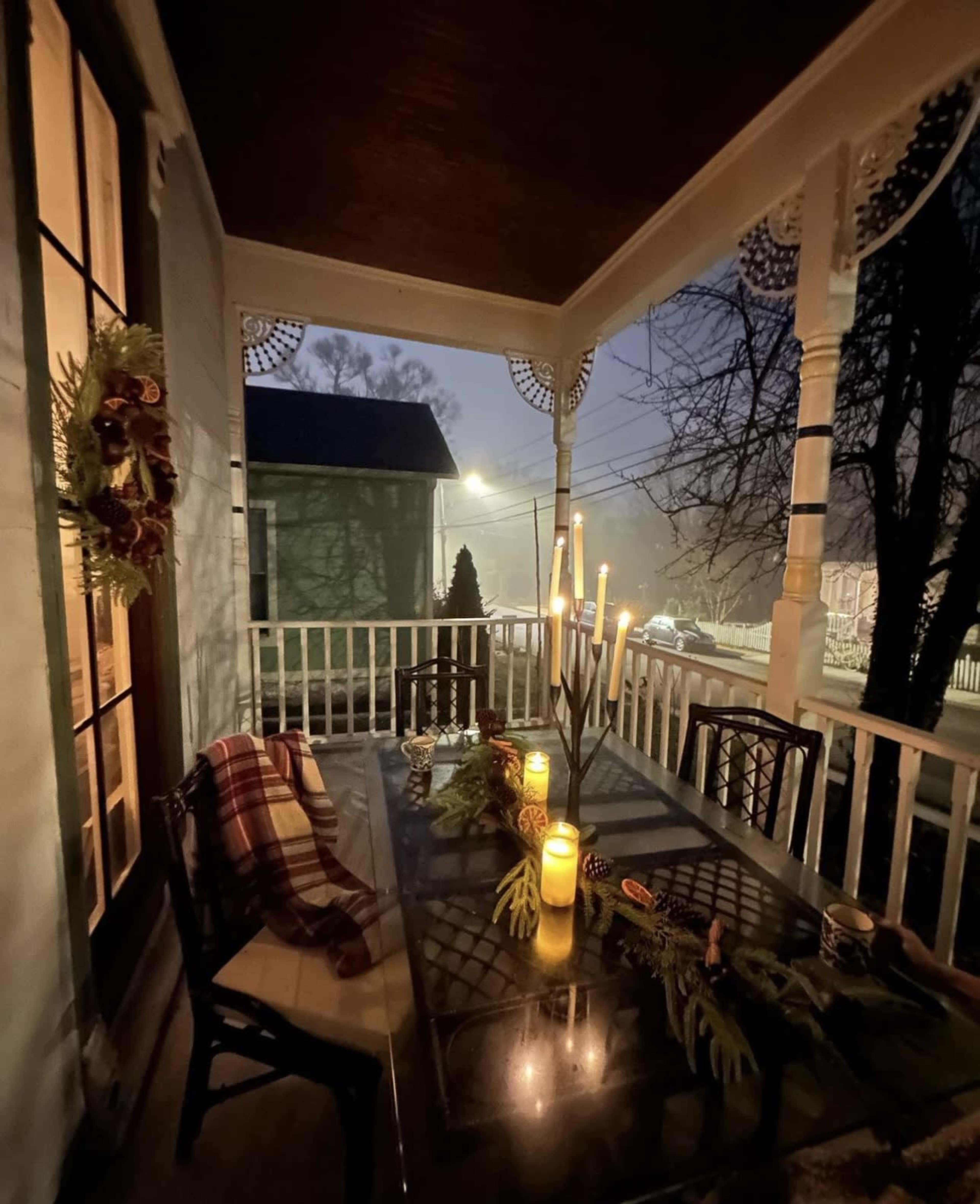The porch is decorated with a green wreath and features a table set for dining with candles and a winter floral arrangement, illuminated by soft light in a foggy atmosphere.