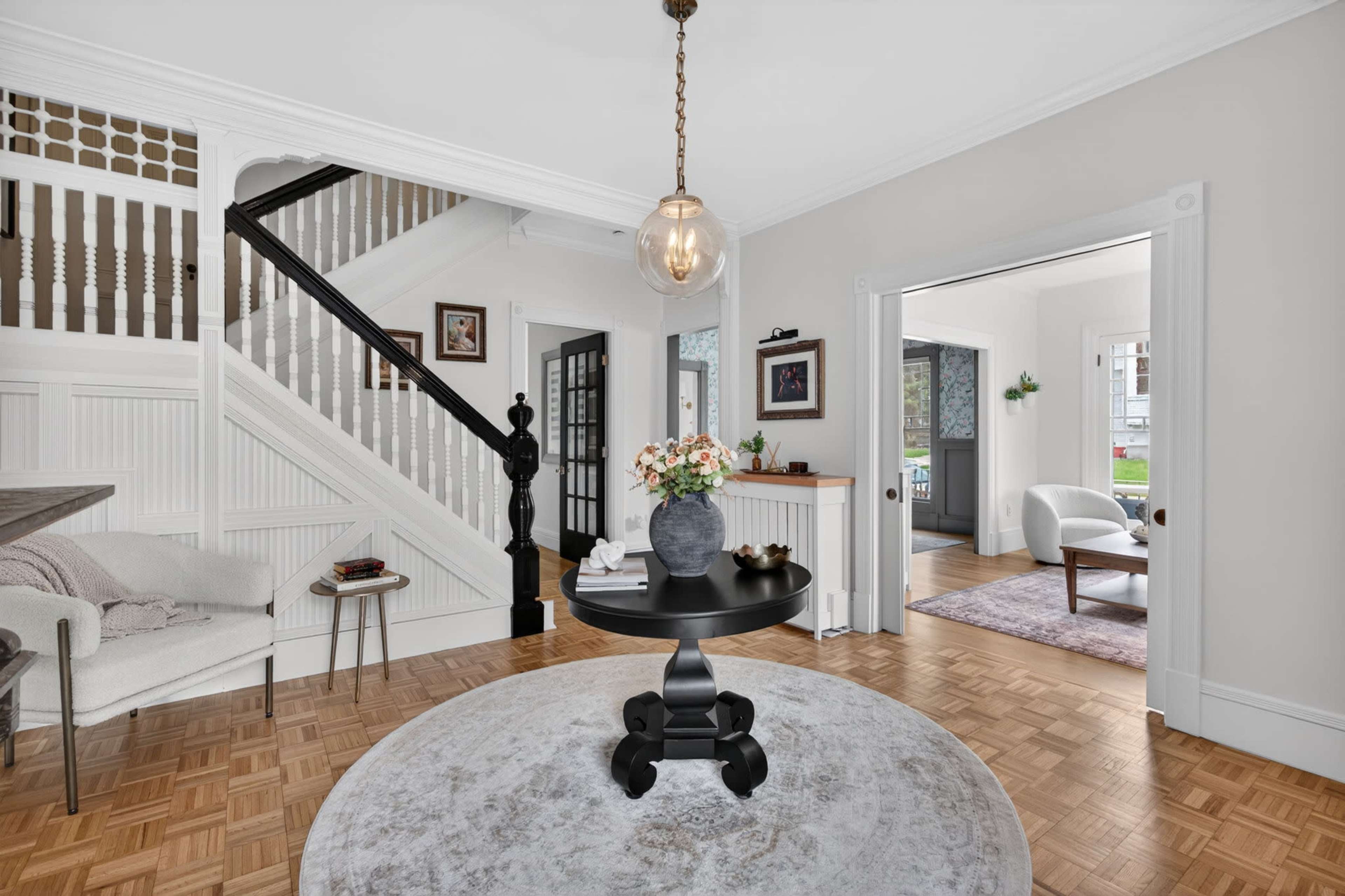 The image shows a well-lit foyer with a round table in the center, a decorative vase on top, and a staircase in the background.