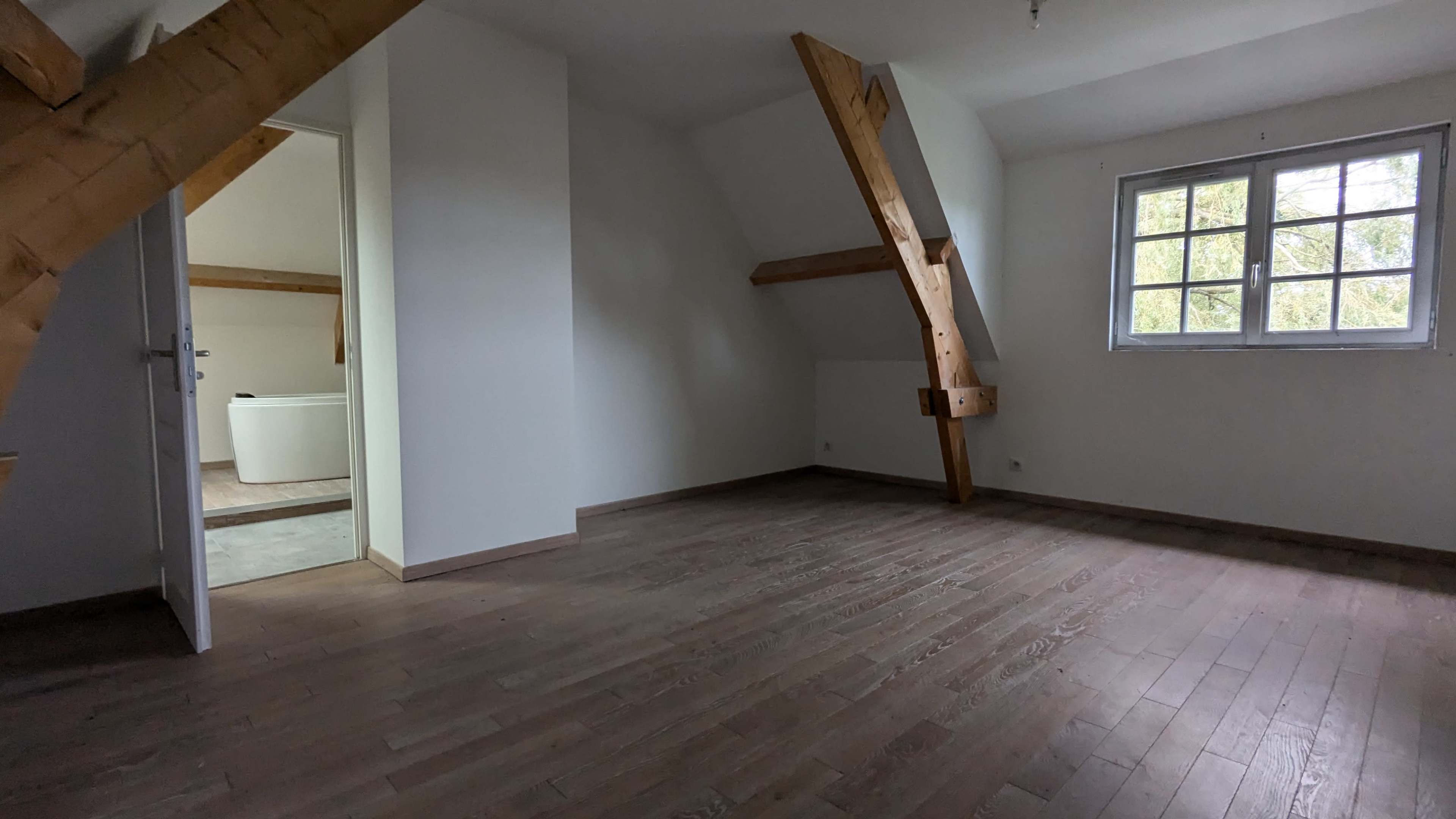 The image shows an empty room with wooden floors, exposed wooden beams, and a doorway leading to a bathroom.
