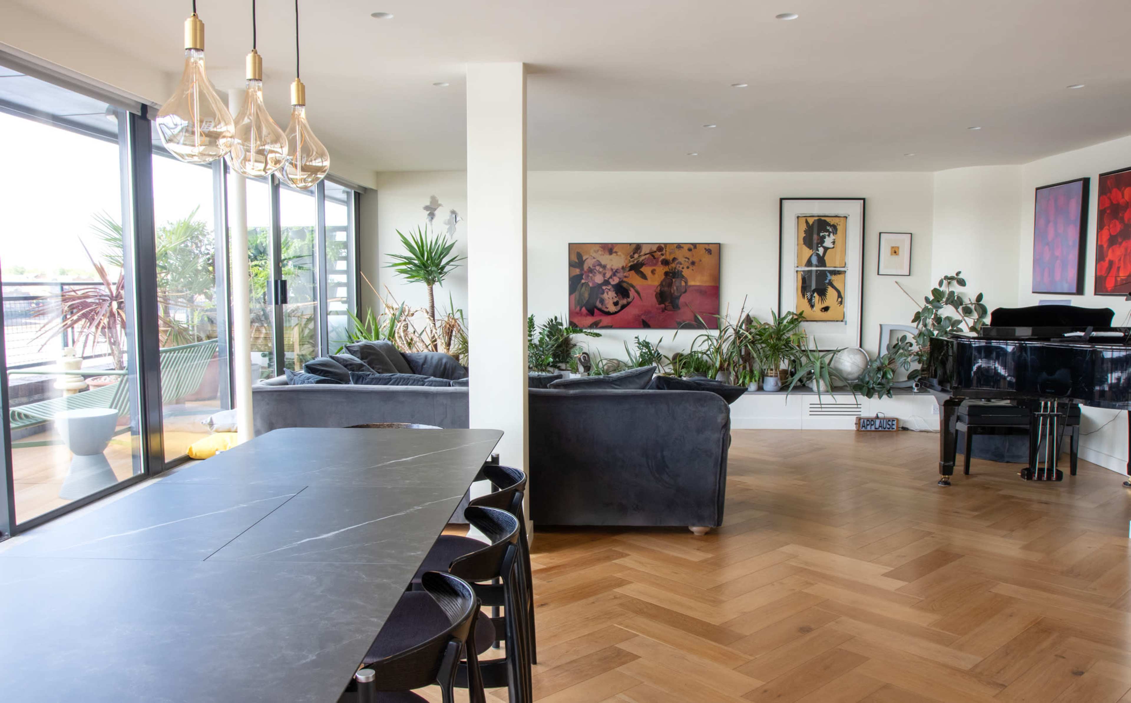 The image depicts a modern living space featuring a large open area with a dining table, two dark sofas, a piano, and numerous plants, all illuminated by natural light through large windows.