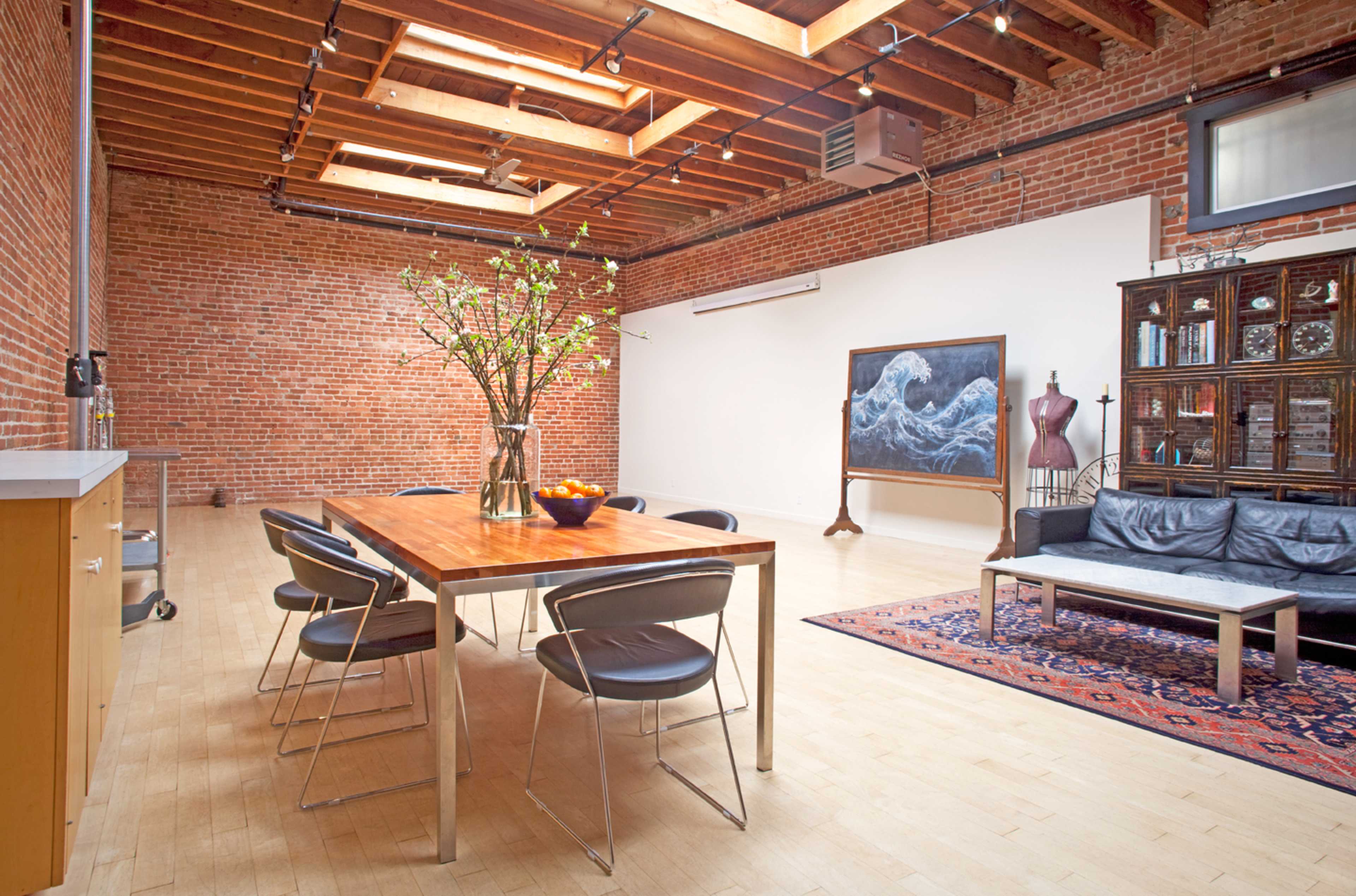 A spacious room with exposed brick walls, featuring a dining table with chairs, a floral centerpiece, and a sofa opposite a large decorative painting.