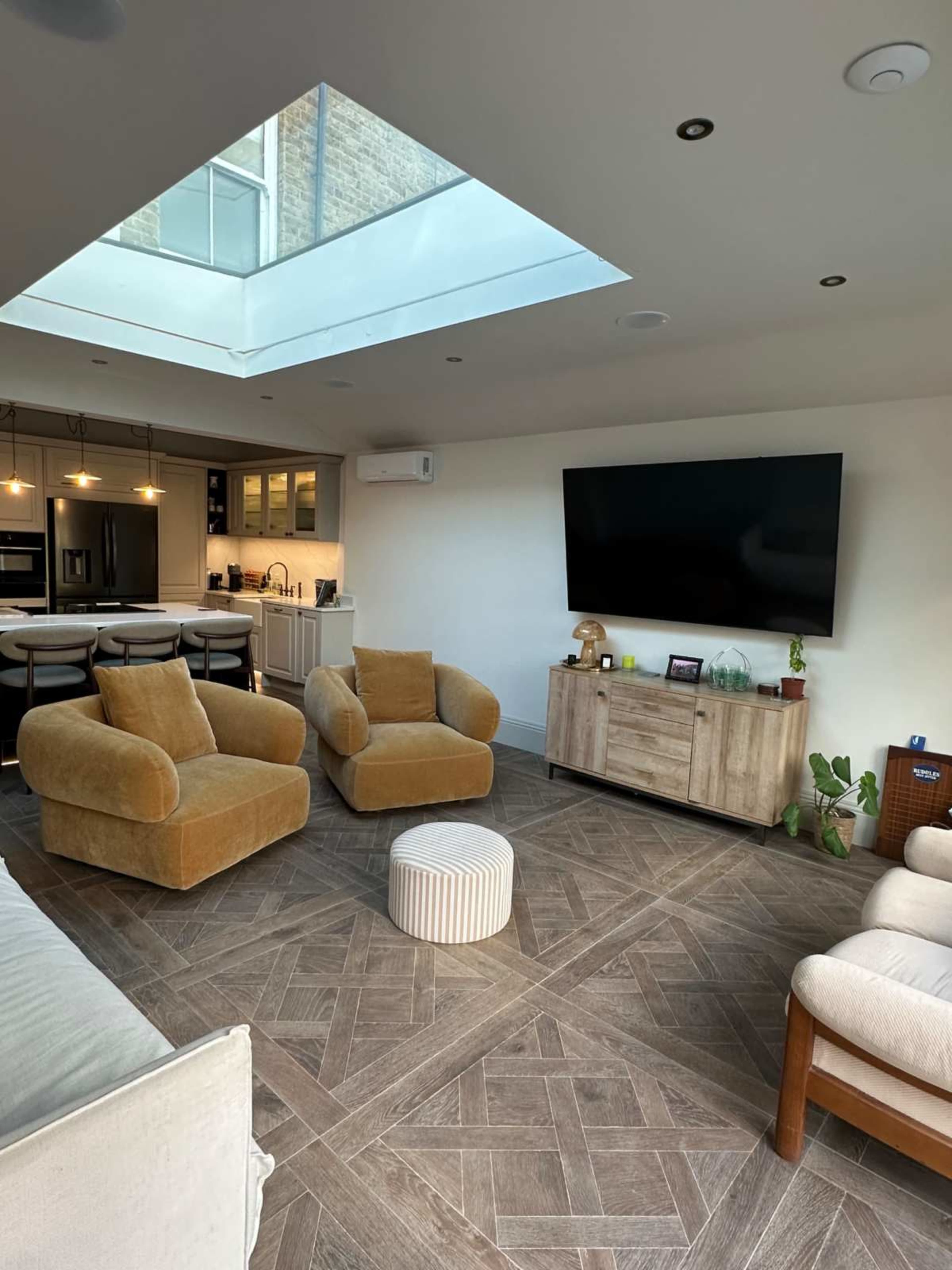 The image shows a modern living room with two brown armchairs, a light-colored pouf, a wooden TV stand, and a skylight above, leading into an adjoining kitchen area.