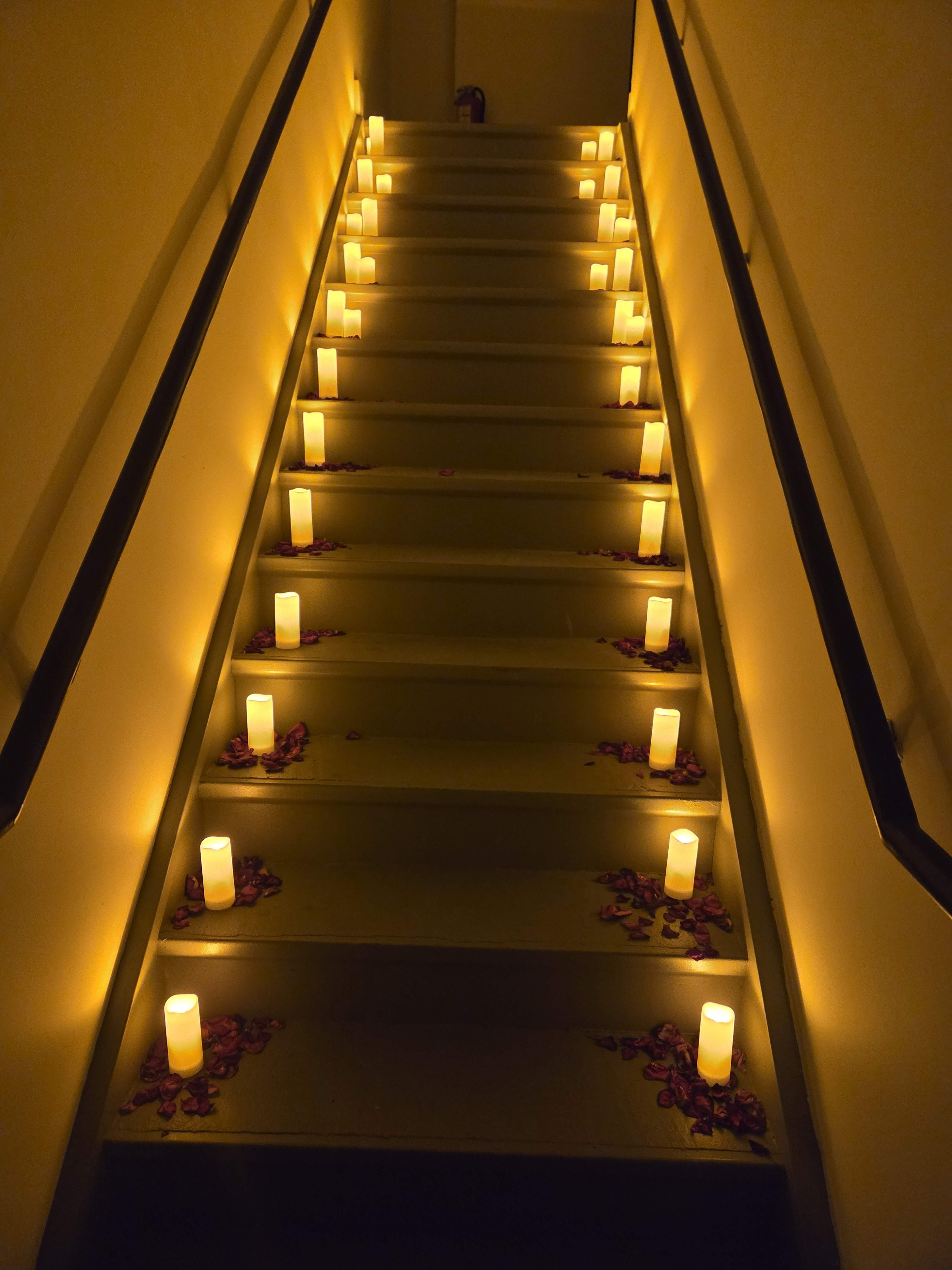 A staircase is lined with illuminated candles and scattered rose petals, creating a warm ambiance.