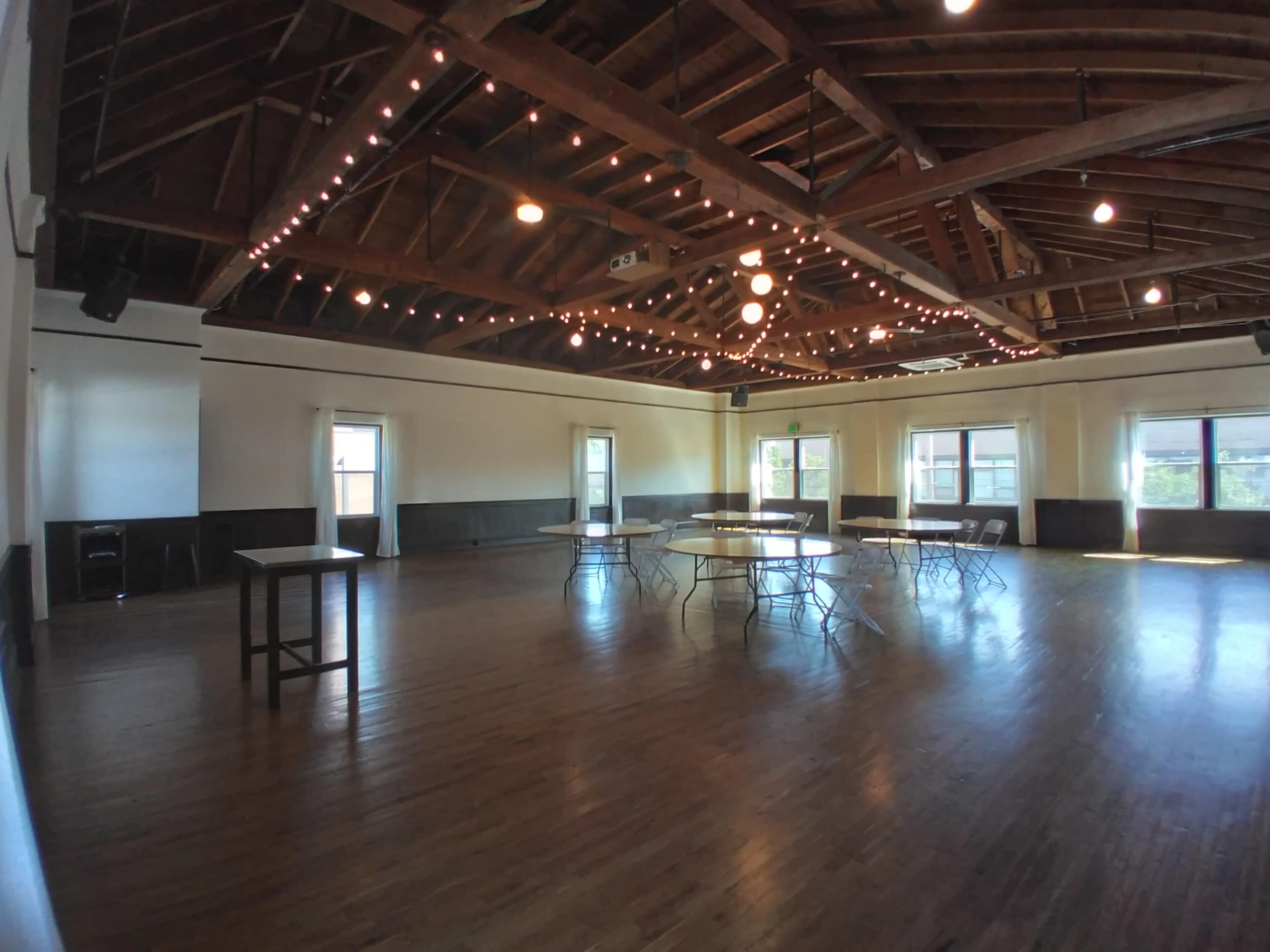 A spacious event venue with wooden floors, exposed beams, and string lights overhead, featuring several round tables set up around the room.