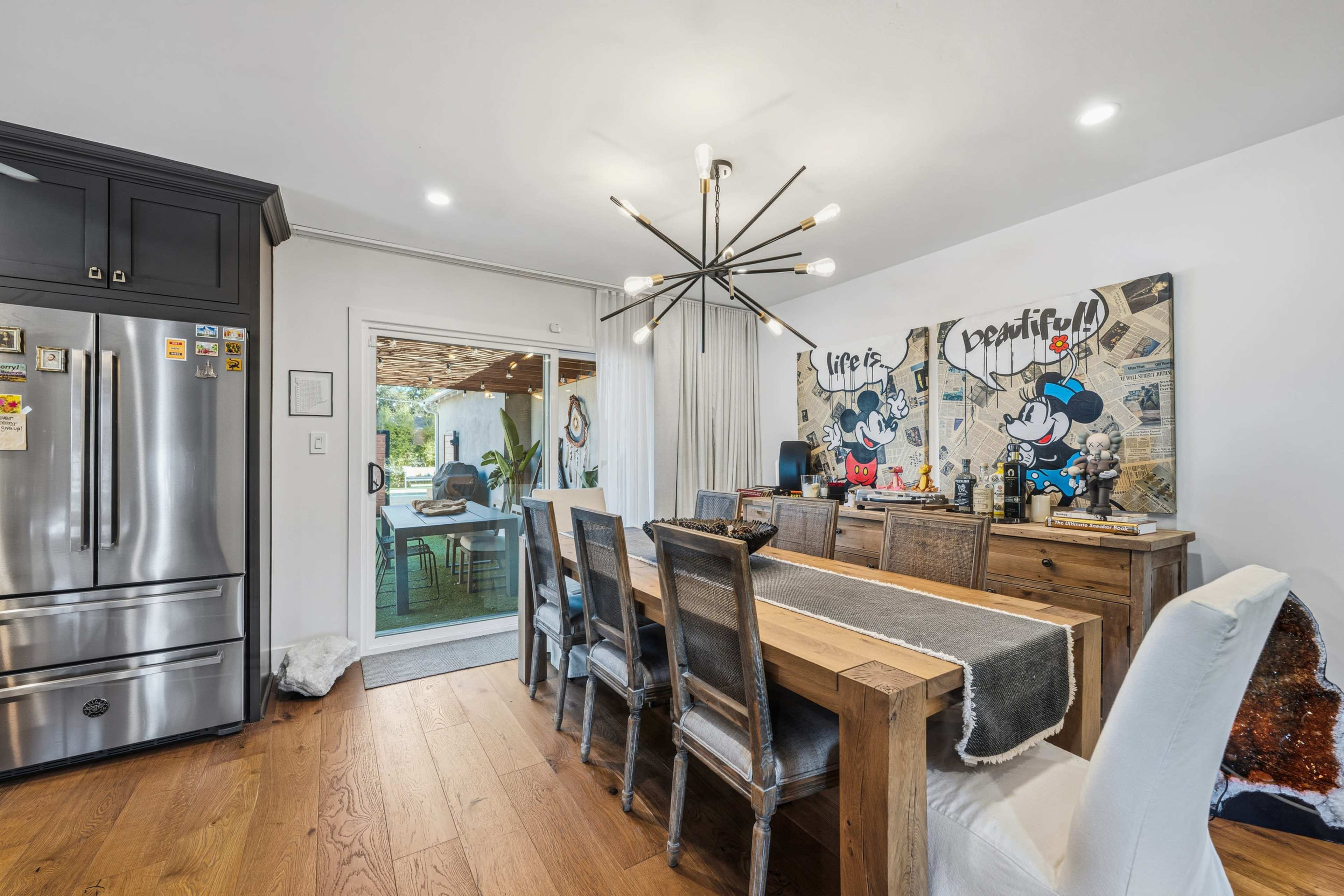 The dining area features a long wooden table surrounded by chairs, with modern artwork on the walls and a large sliding glass door leading to an outdoor space.