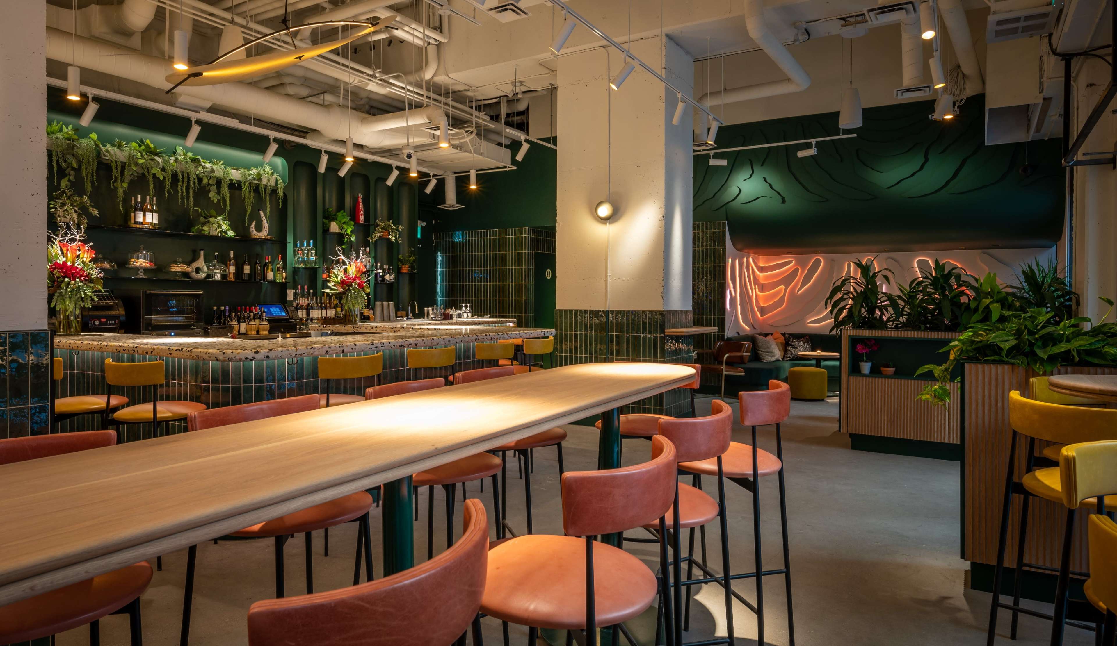 The image shows a modern bar area with a long wooden countertop, colorful stools, and green foliage decorating the walls and shelves.