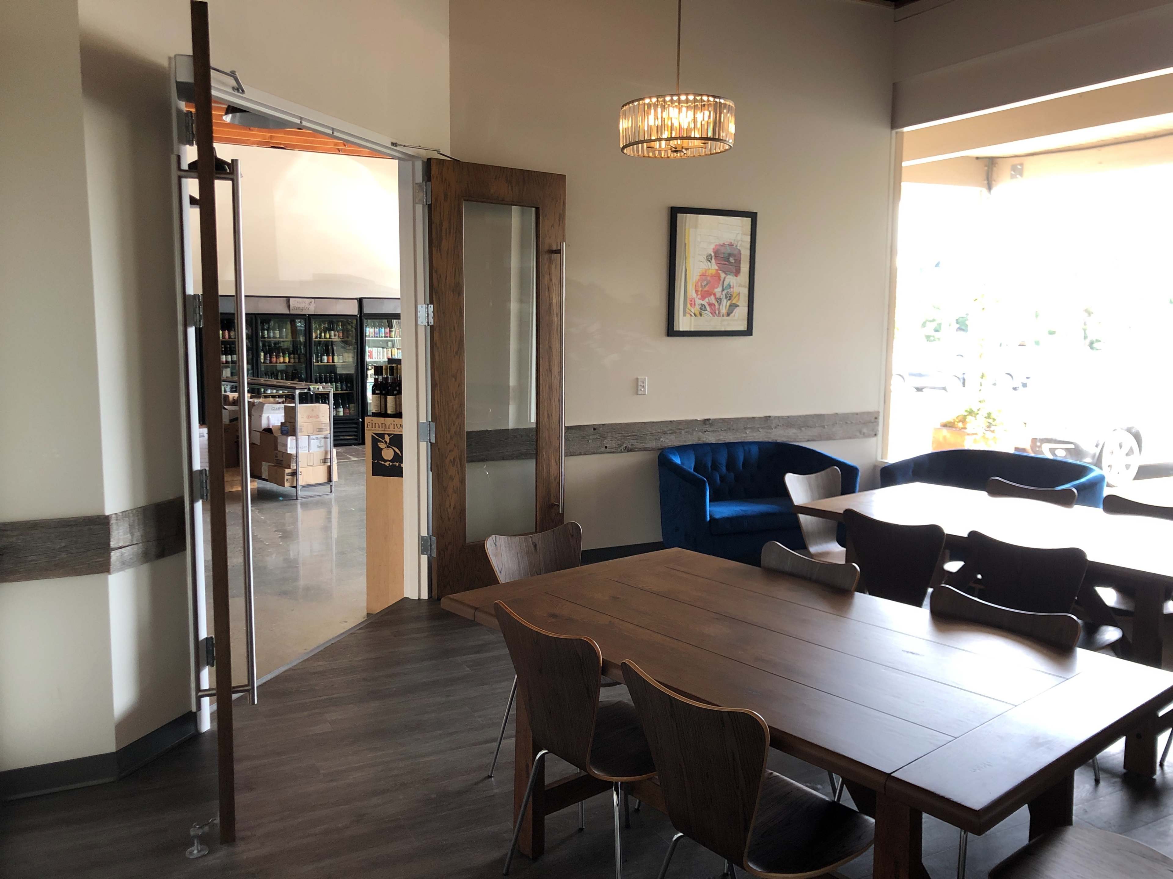 A modern interior of a cafe or restaurant with a wooden dining table, blue chairs, and a view of shelving units in an adjacent room.