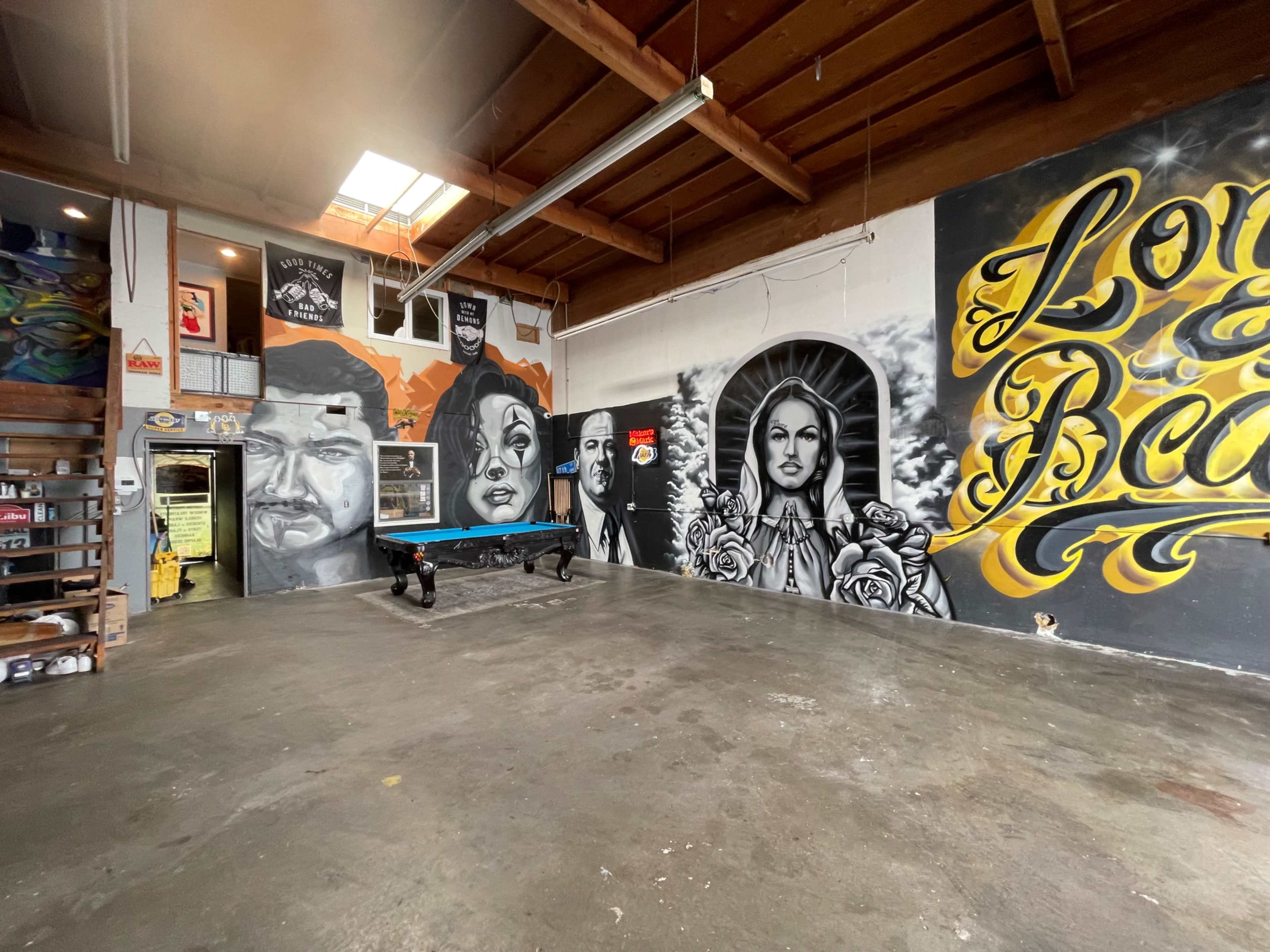 An industrial-style room featuring large, intricate murals on the walls, a pool table in the center, and shelves with various items along one side.