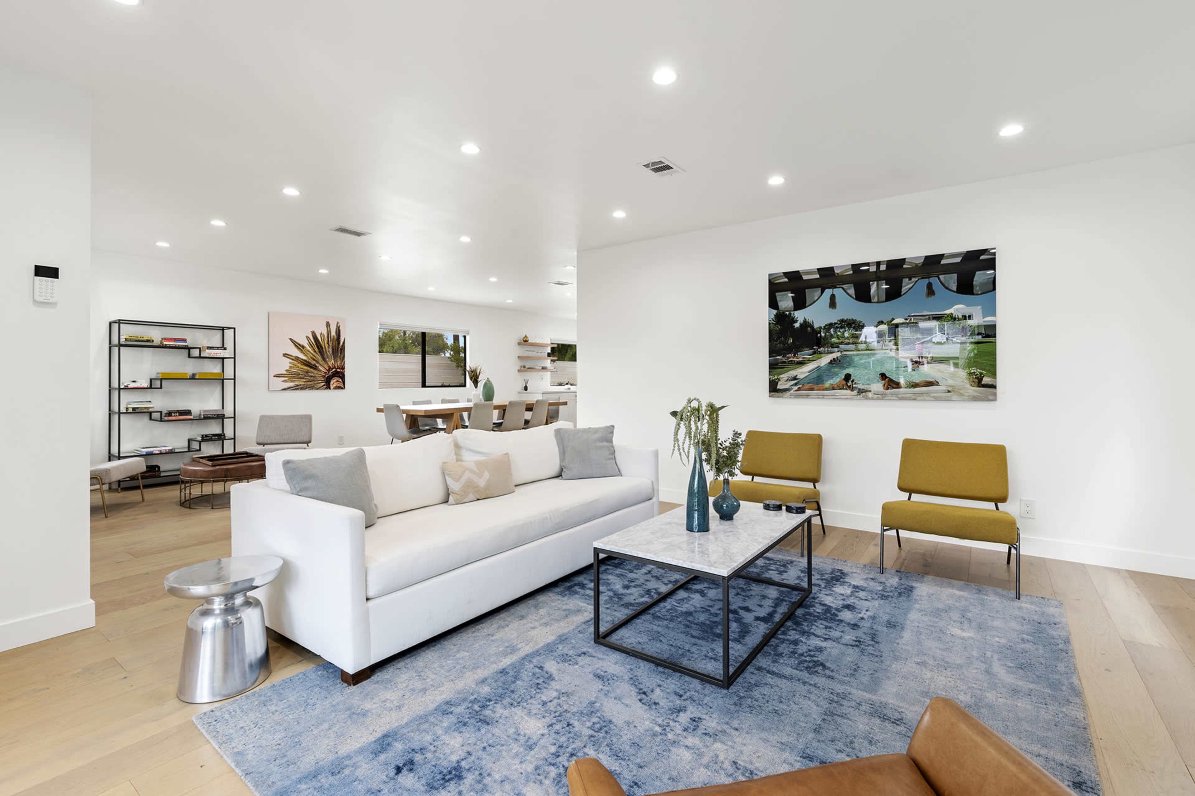 A modern living room with a white sofa, two mustard-colored chairs, a coffee table, and a blue rug, featuring a large artwork on the wall.