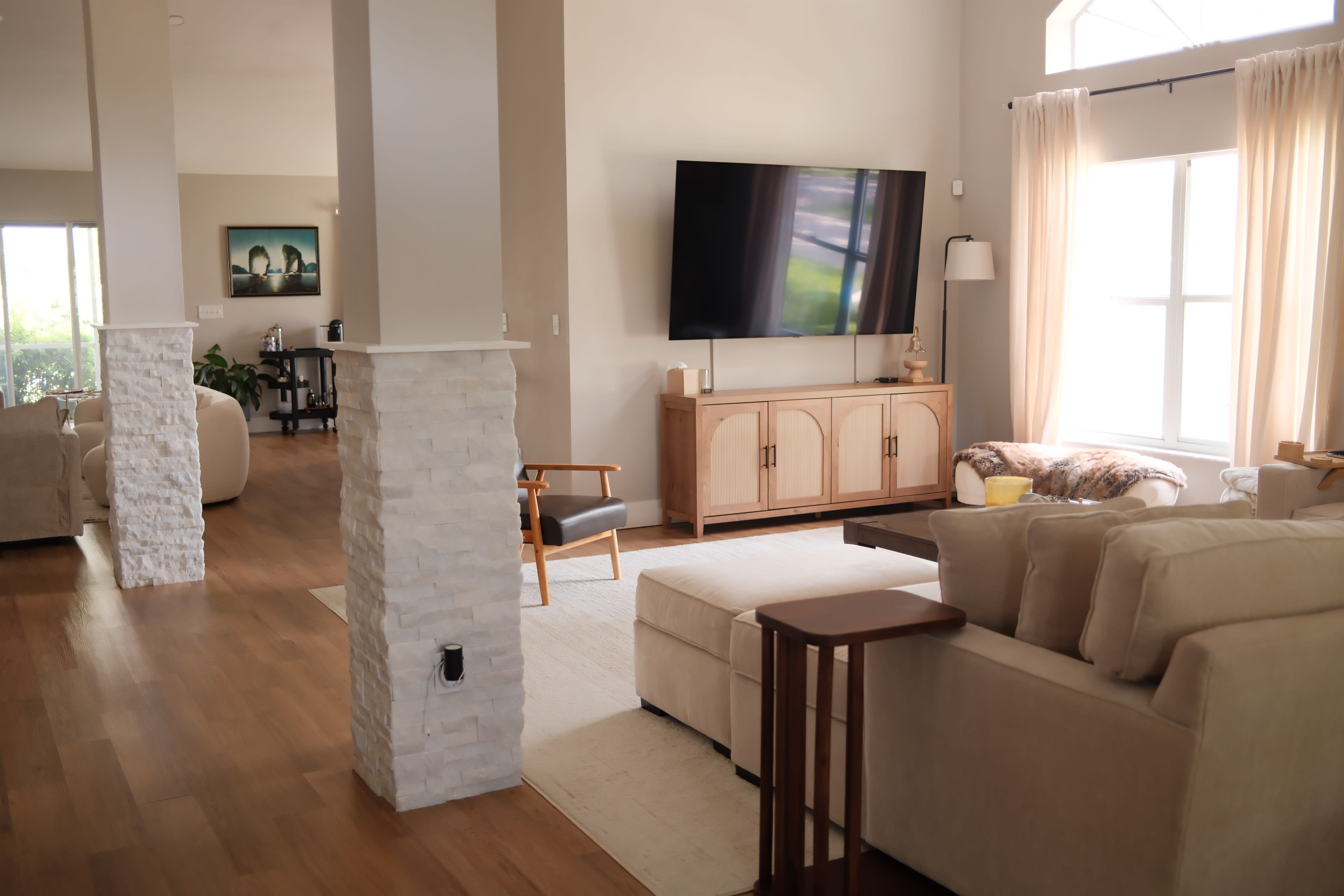 The image shows a modern living room with a large television on the wall, a beige sofa, and a light wooden console table, complemented by natural light filtering through large windows.