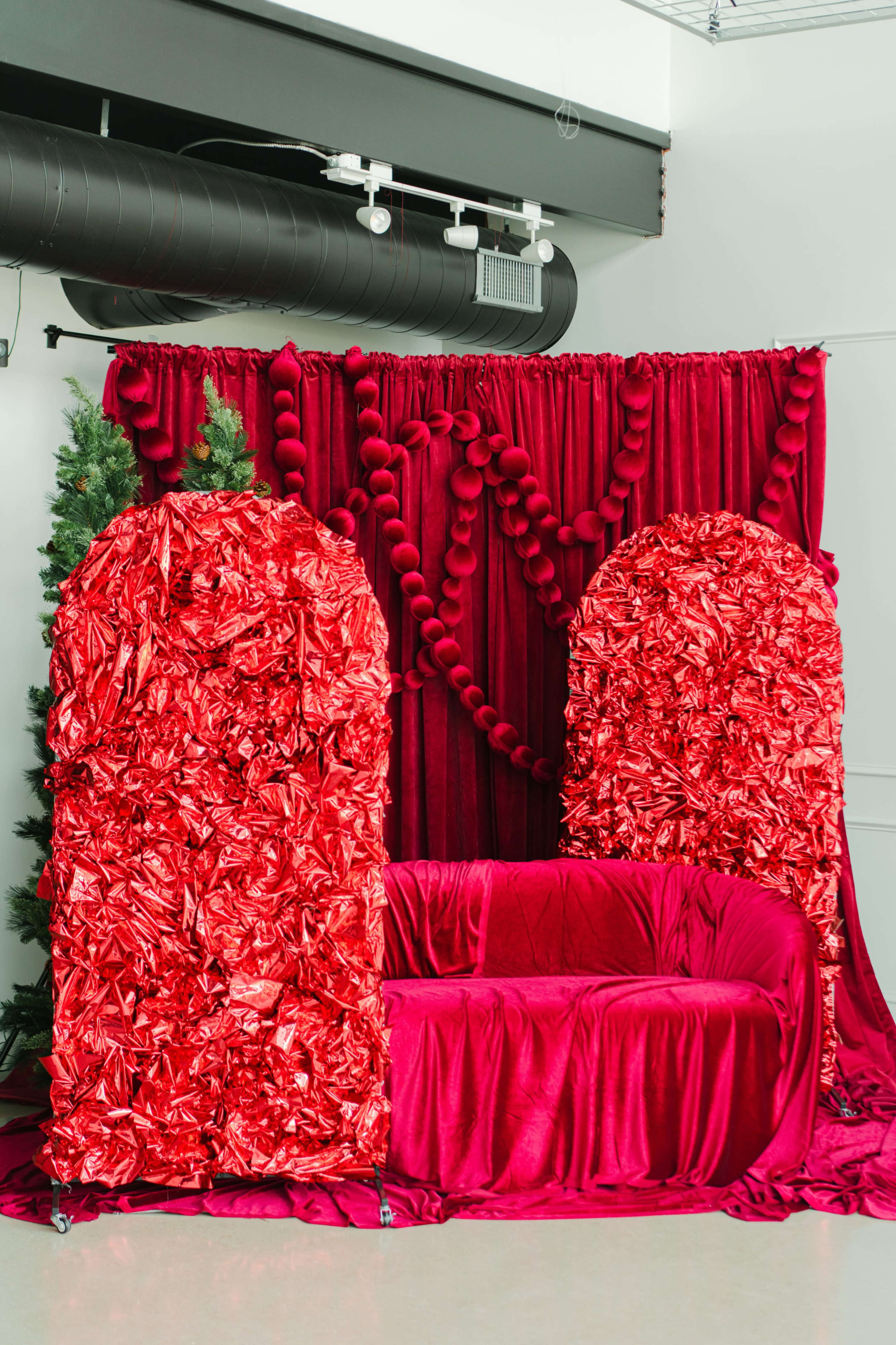 The image features a red velvet sofa surrounded by two large, textured red decor pieces and a backdrop adorned with red drapery and pom-pom accents.