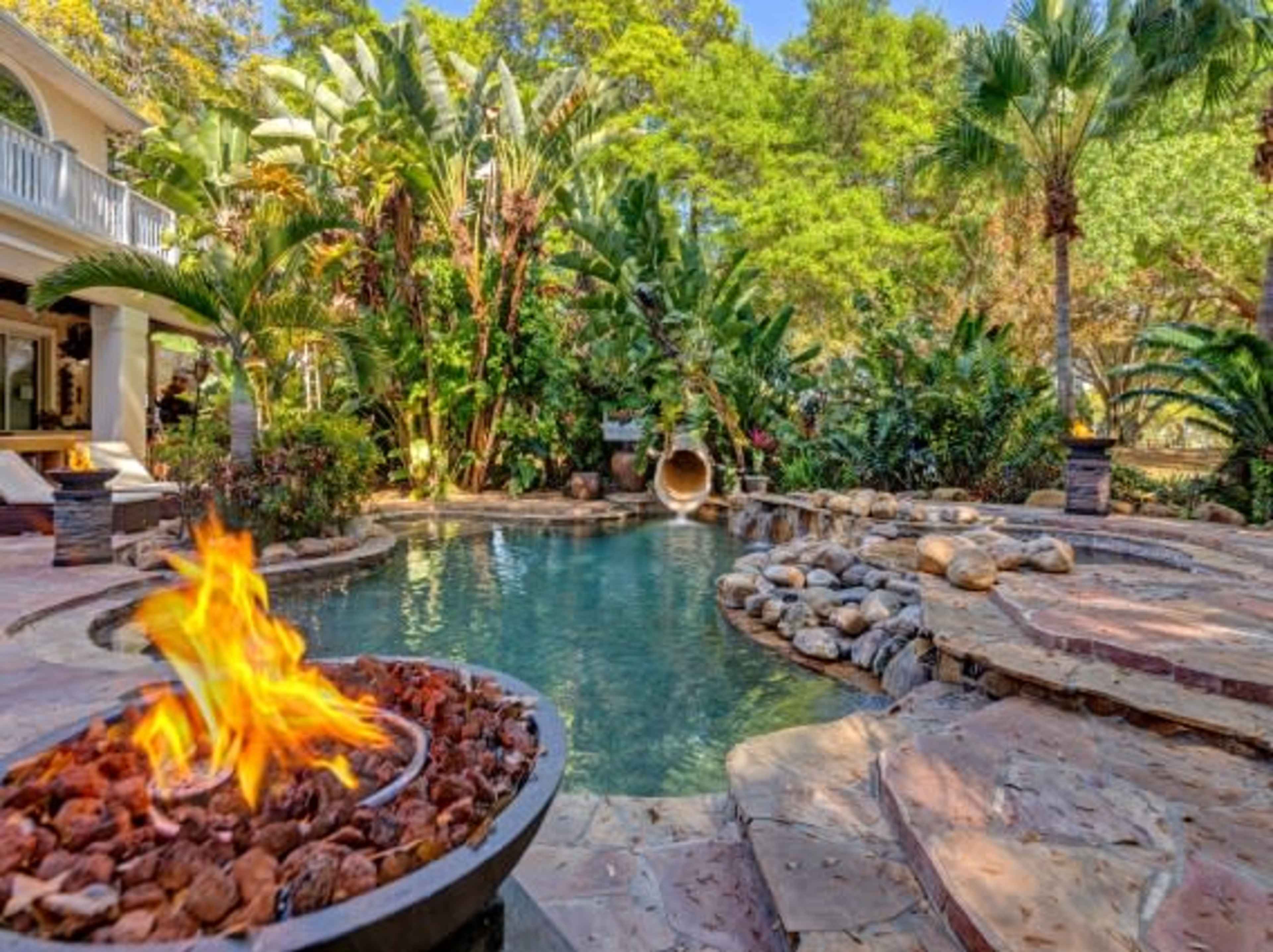 A serene backyard with a swimming pool, a stone patio, tropical plants, and a fire pit.