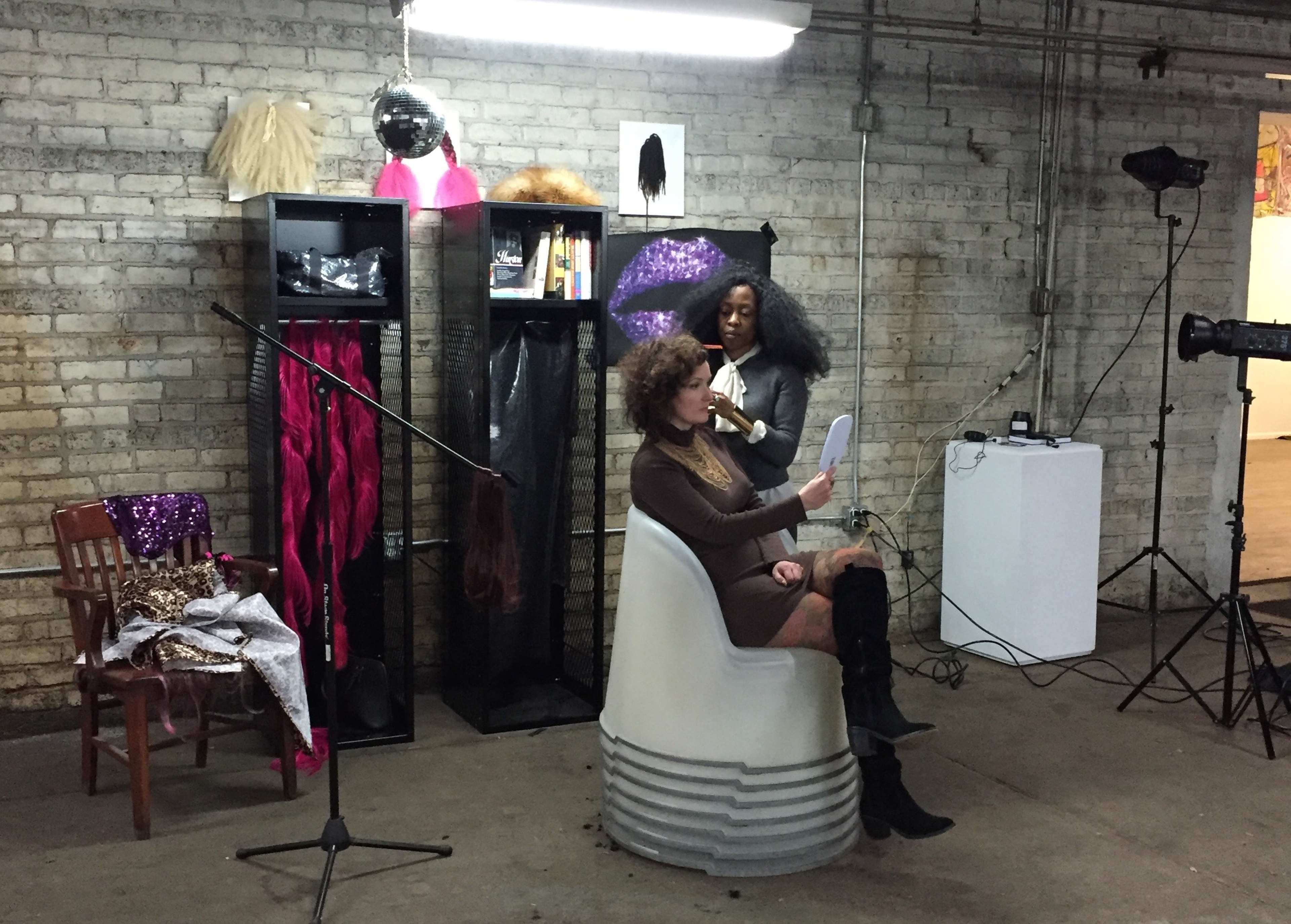 A woman sits in a chair holding a mirror, while another woman stands nearby in a space featuring hair displays and a microphone setup.