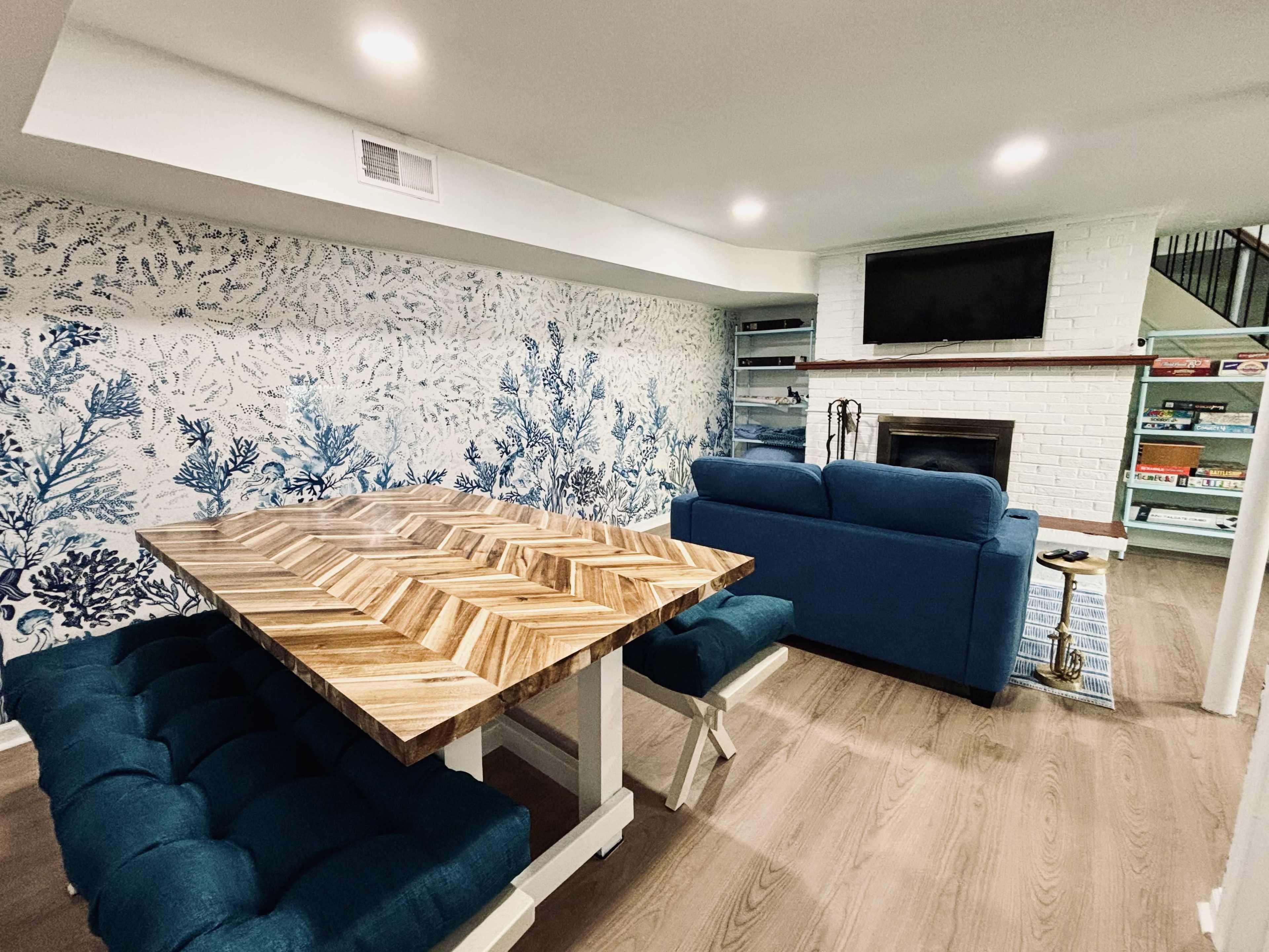A living space features a patterned blue wallpaper, a wooden dining table, a blue couch, and a fireplace with a television mounted above it.