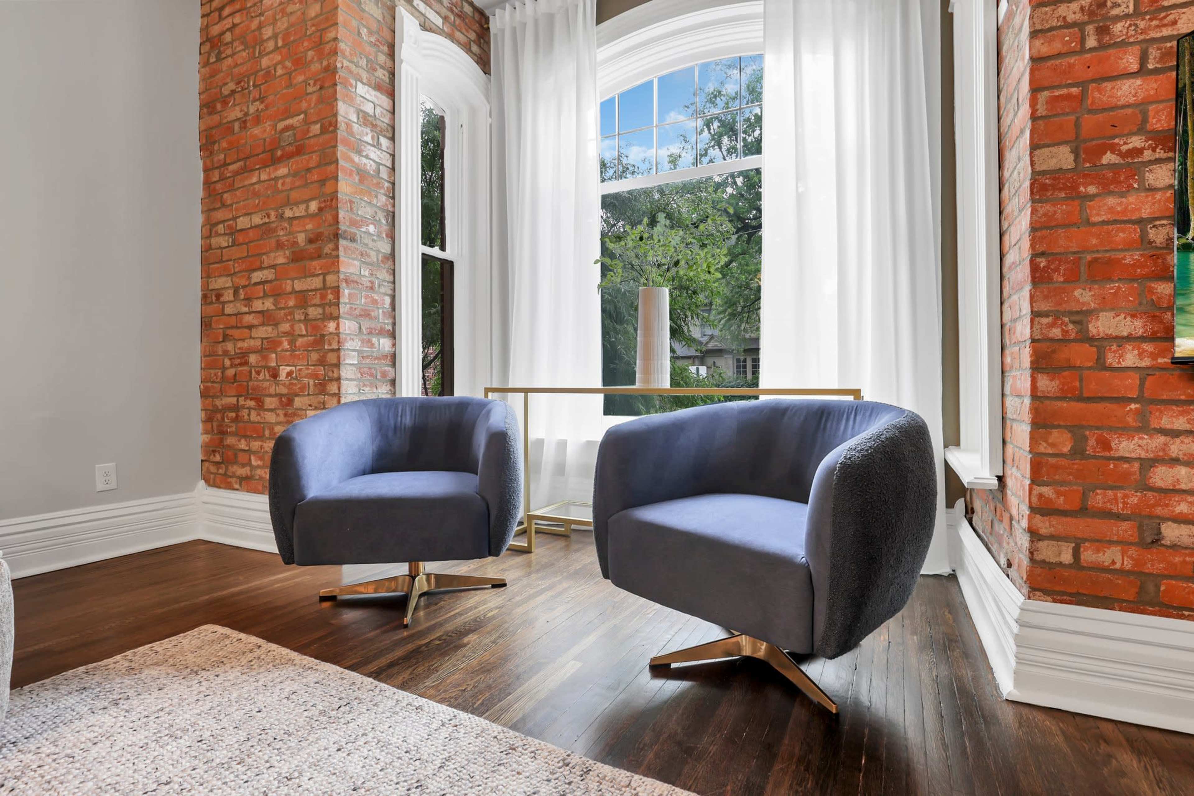 The scene features two modern blue armchairs positioned near a large window, with brick walls and a wooden floor creating a cozy atmosphere.