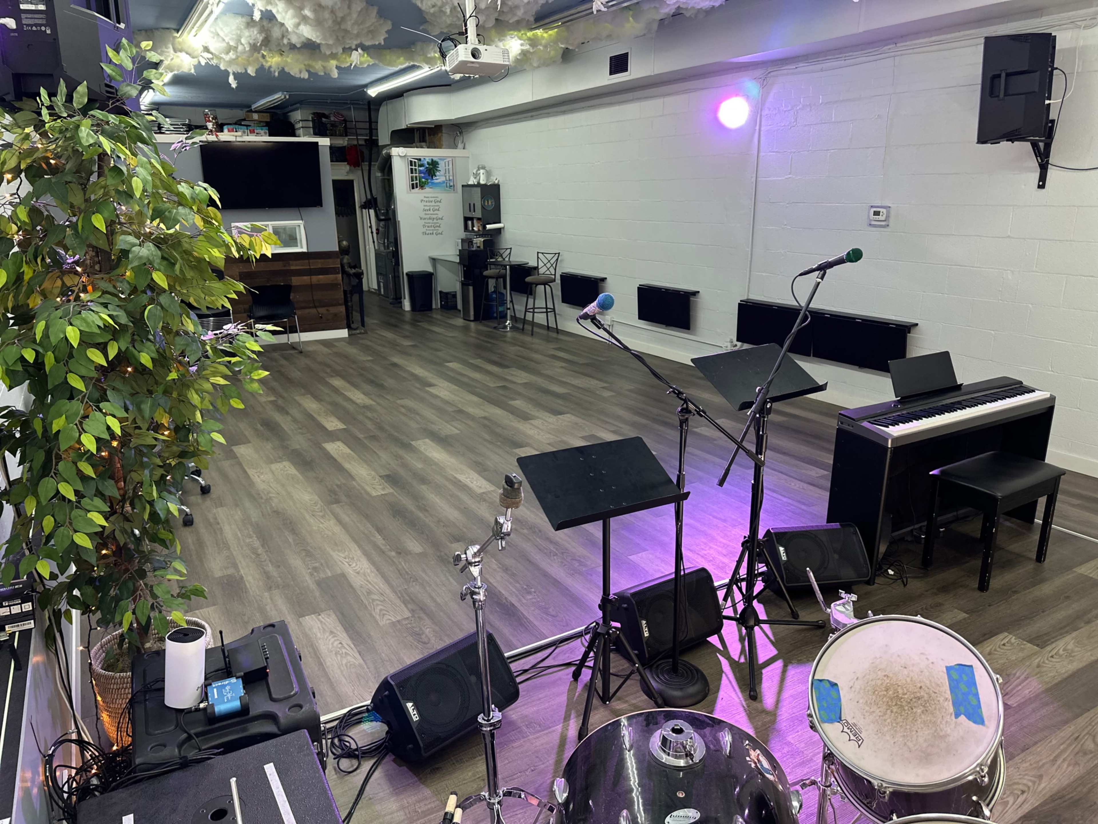 The image shows a performance space featuring musical equipment, including a drum set, a keyboard, and microphones, set in a well-lit room with wooden floors.