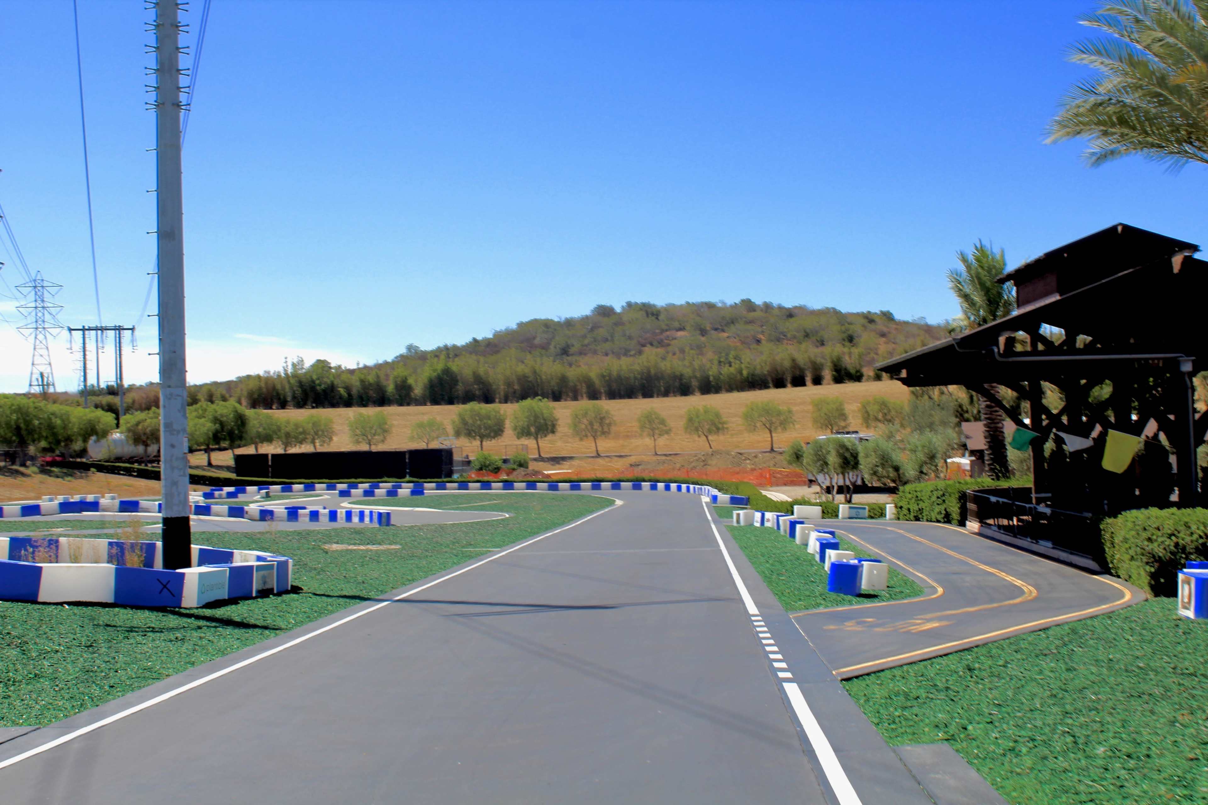 The image shows a racetrack with a winding asphalt path bordered by blue and white barriers, set against a backdrop of green hills and clear blue skies.