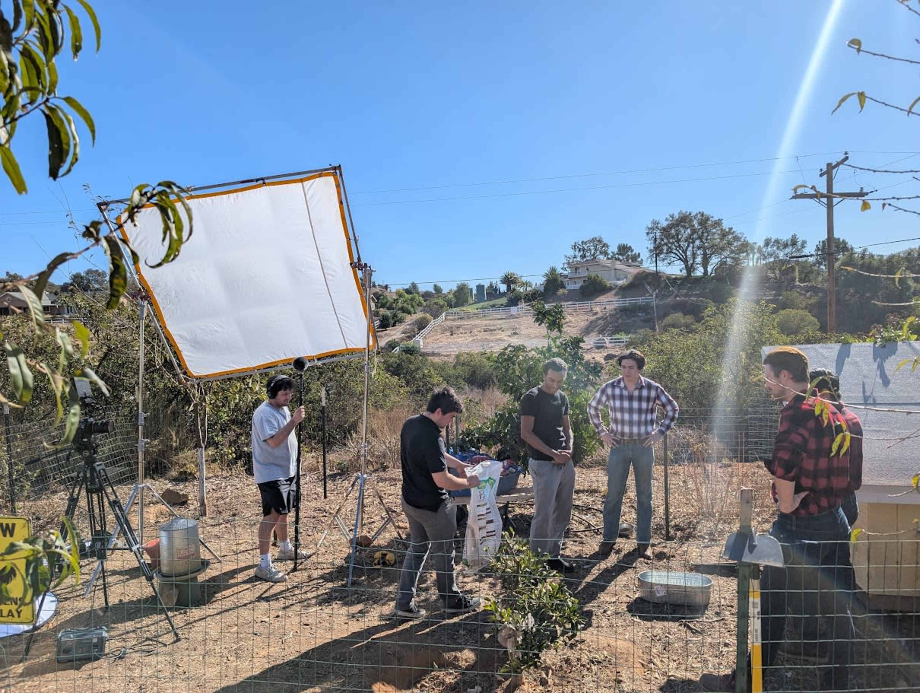 A group of five people is gathered outside in a field setting, with a large white reflective screen and equipment nearby.