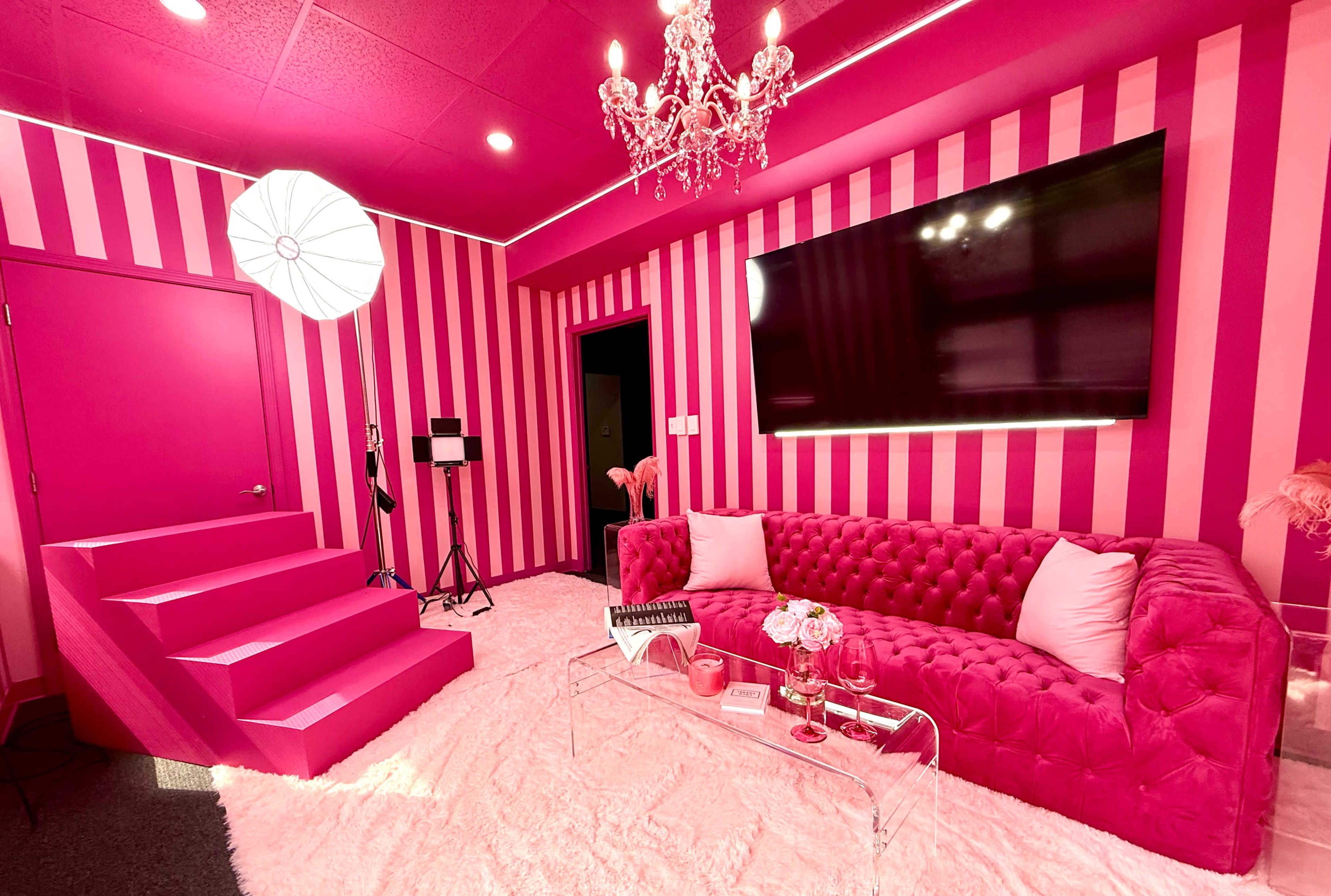 The room features bright pink striped walls, a plush pink couch, a crystal chandelier, and a large television, with a fluffy white rug covering the floor.