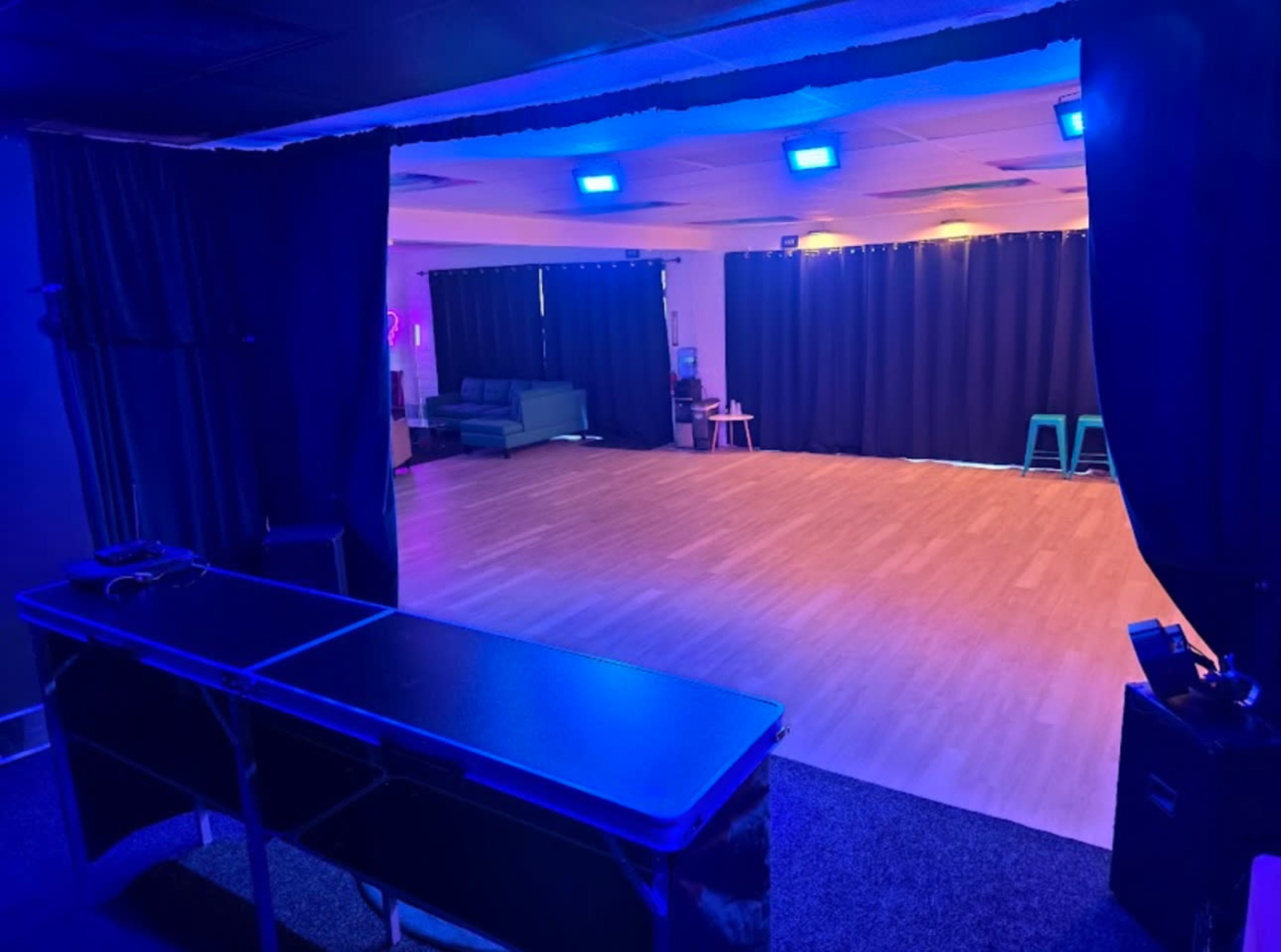 The image shows a spacious room with hardwood flooring and dim lighting, featuring a seating area and black curtains on one side.