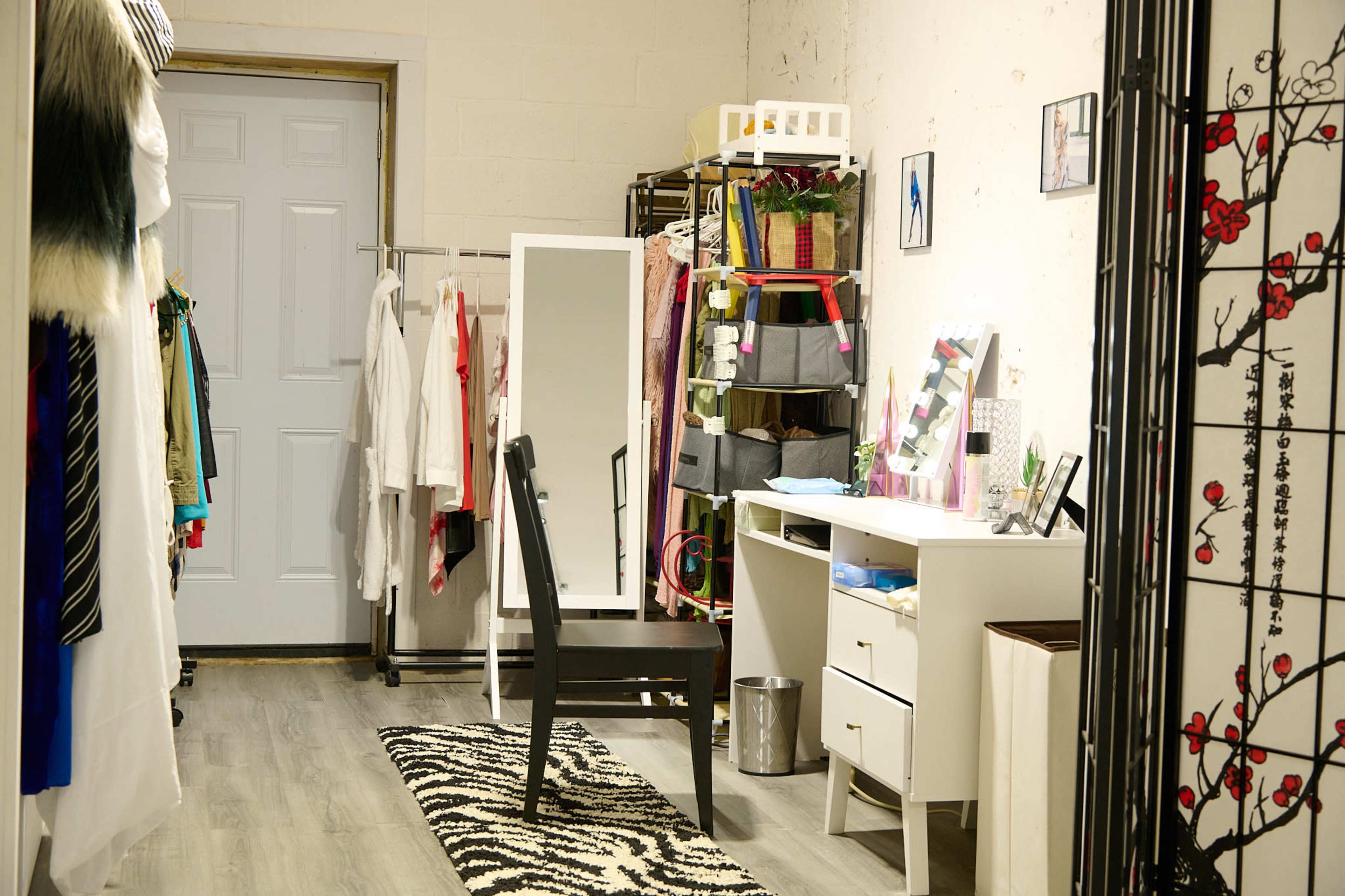 A room with a desk, a chair, and various clothing items organized on racks and shelves.