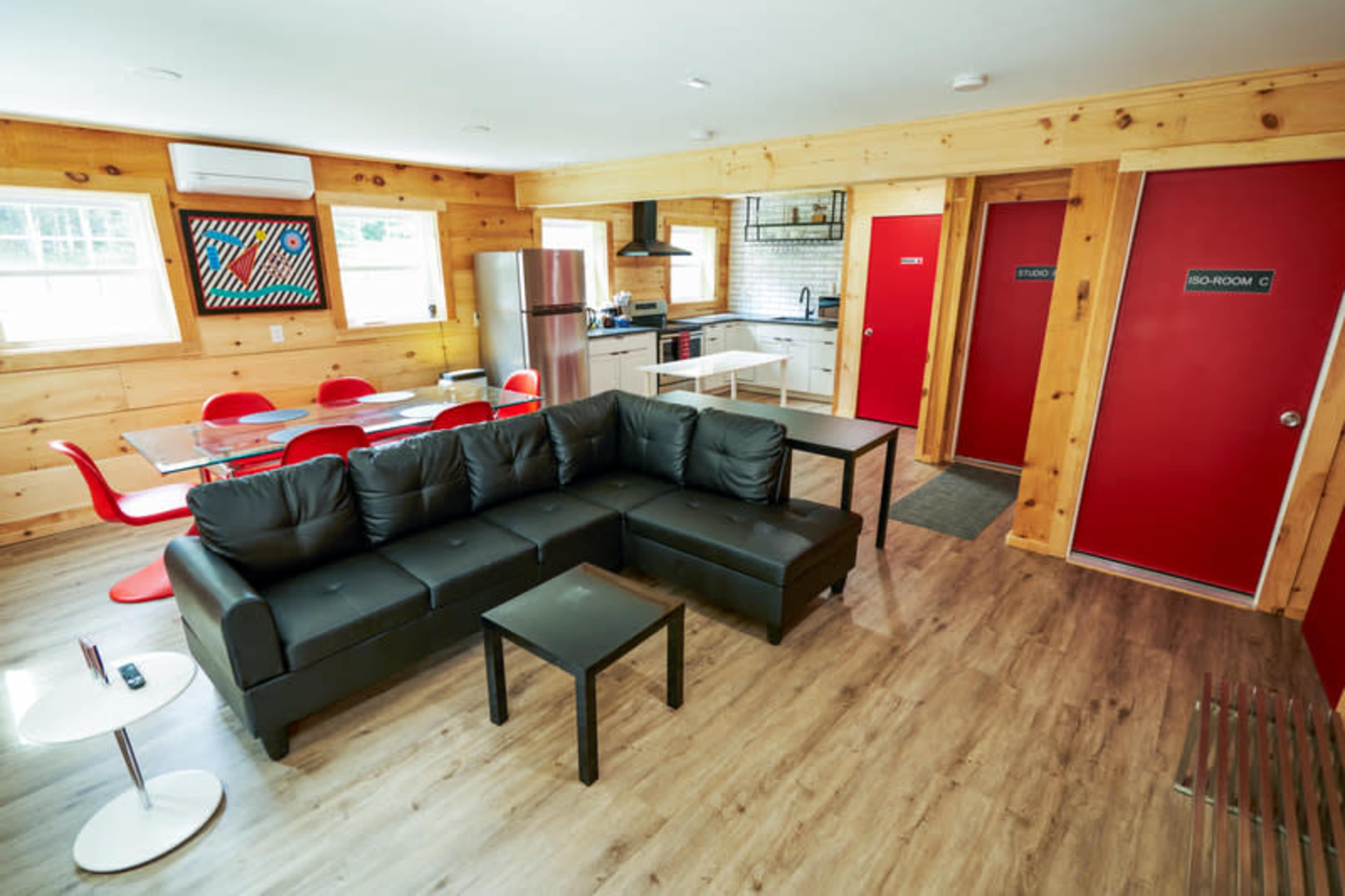 A modern interior of a cabin living space featuring a black sectional sofa, a dining area with red chairs, and a kitchenette with stainless steel appliances.