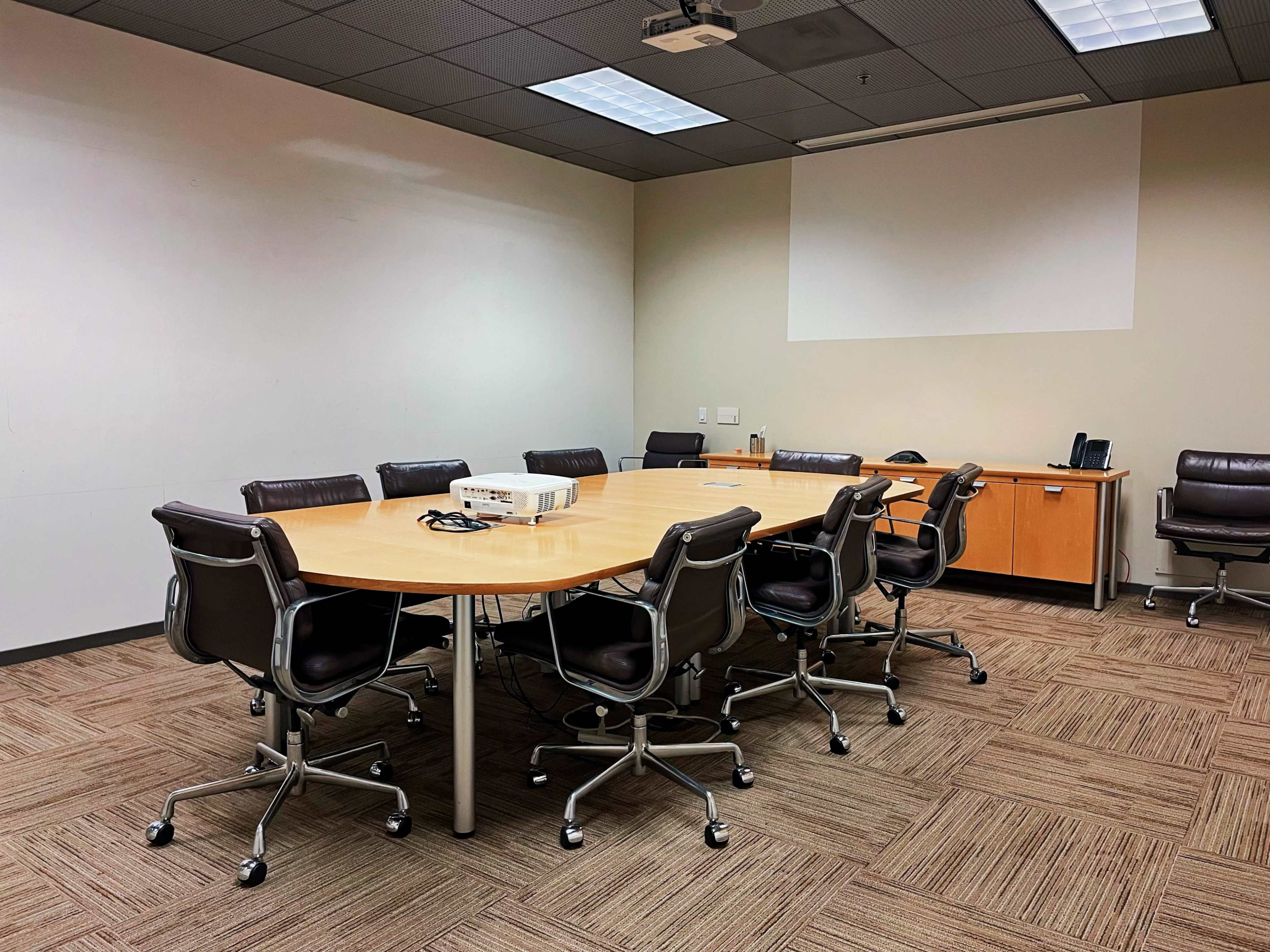 A conference room features a large round table surrounded by several ergonomic chairs, with a projector and a media console positioned at one end.