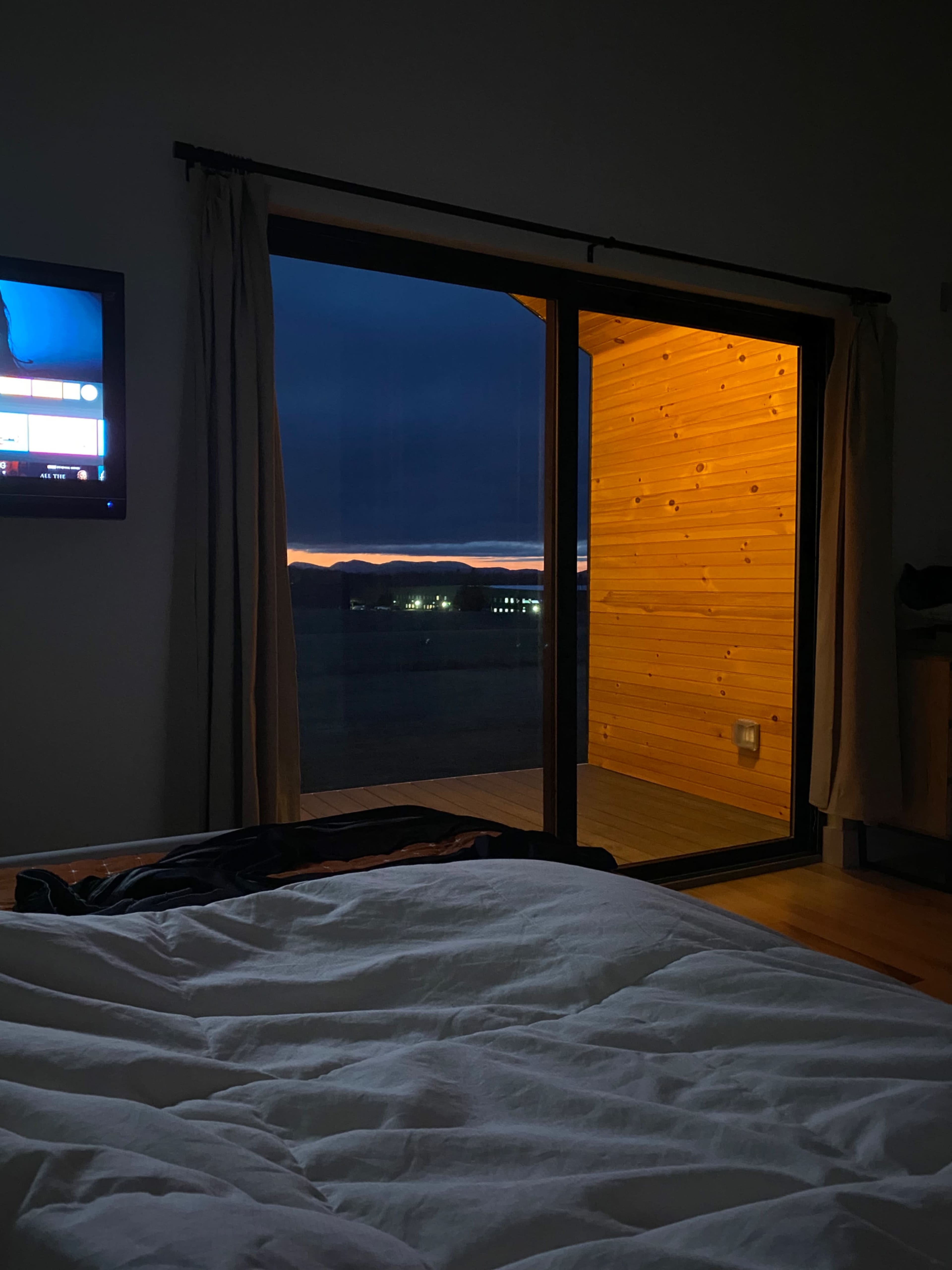 A bedroom scene displays a bed with a white comforter in the foreground, while large glass doors reveal a dimly lit porch and a sunset beyond.