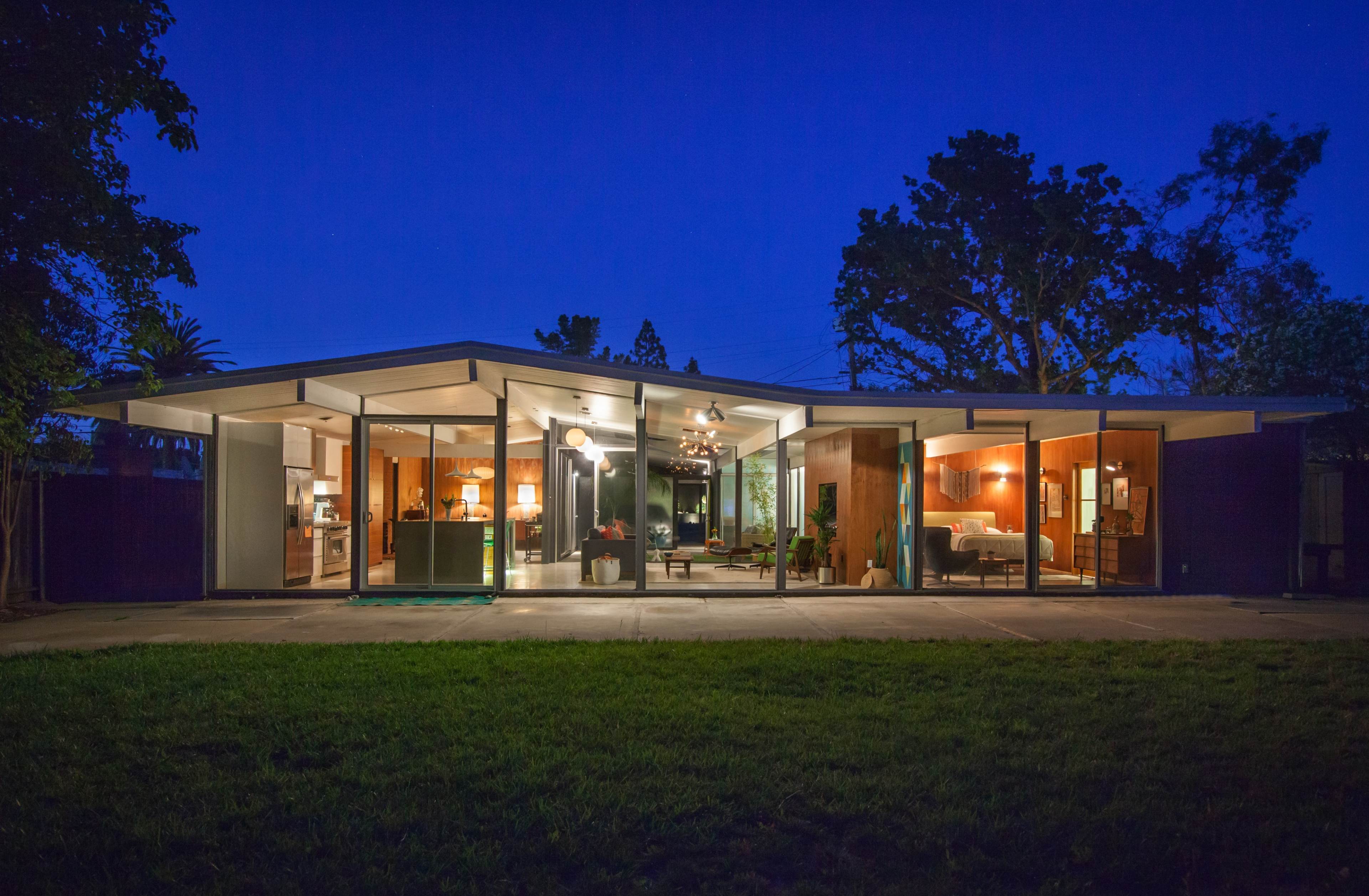 A modern, single-story house with large glass windows is illuminated at night, revealing its interior layout and furnishings.