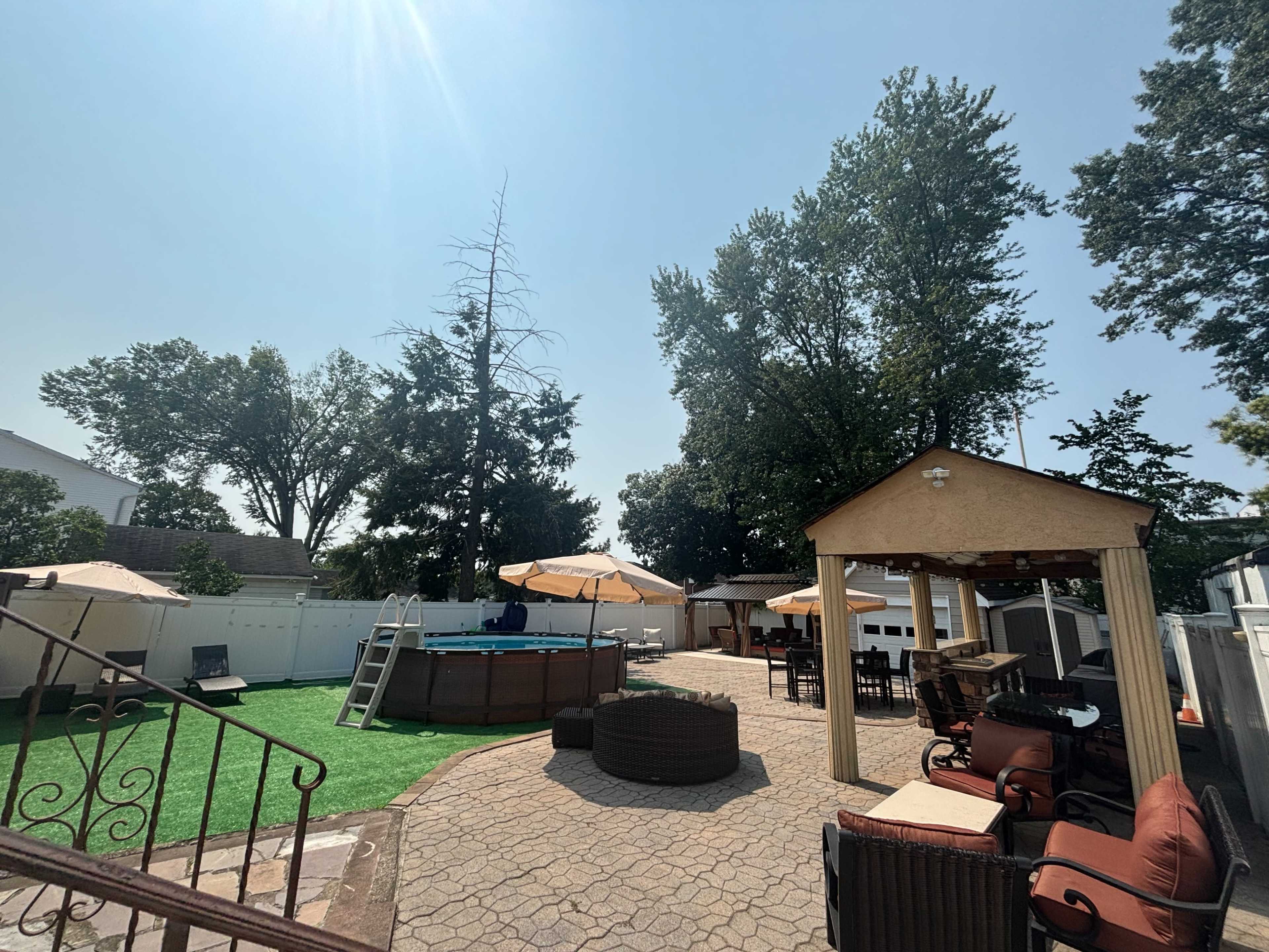 The image shows a backyard with a circular above-ground pool, multiple lounge chairs, outdoor dining furniture, and shade umbrellas surrounded by trees and a fence.