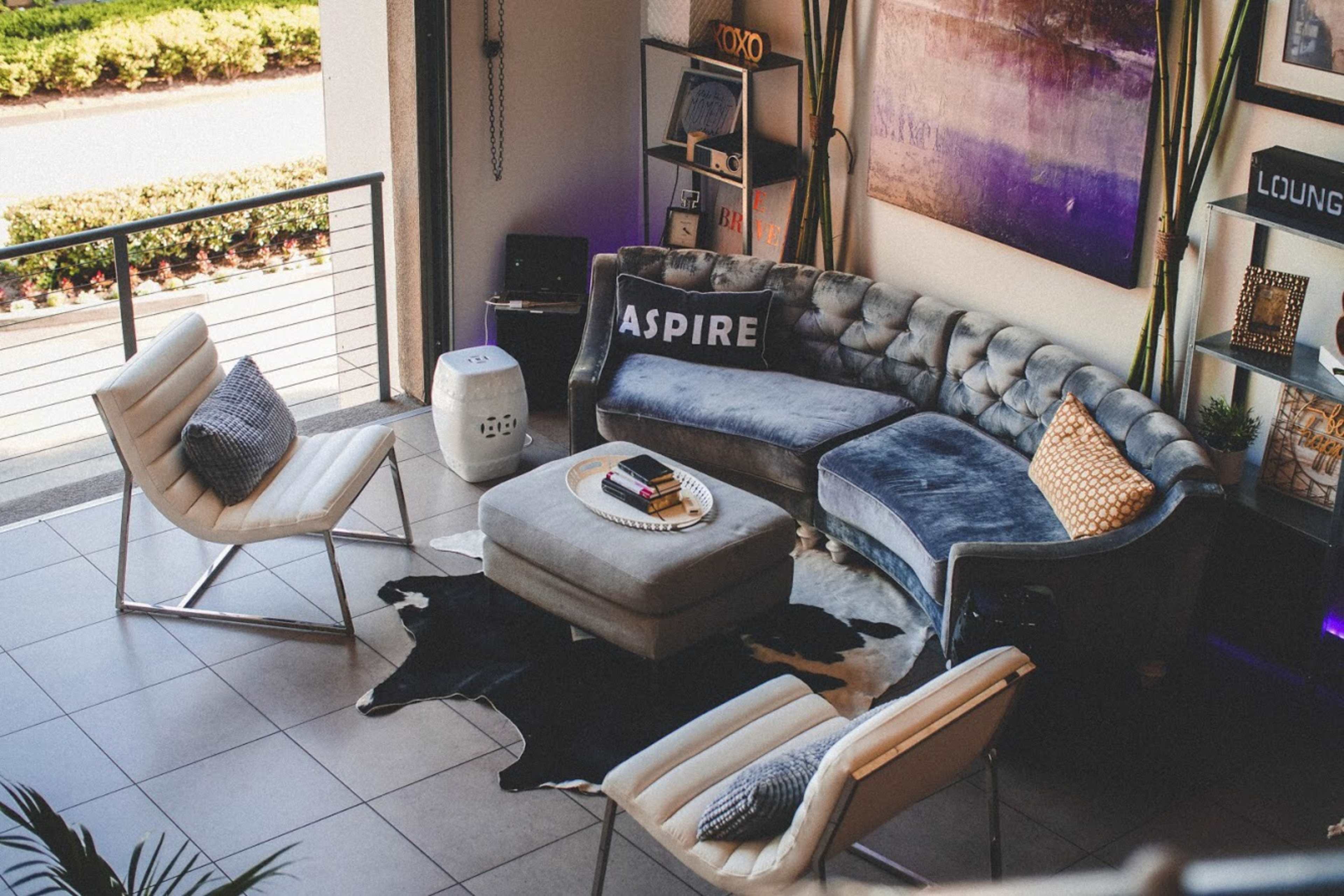 The image shows a modern living room with a tufted blue sectional sofa, two white chairs, a round coffee table, and a black and white cowhide rug.