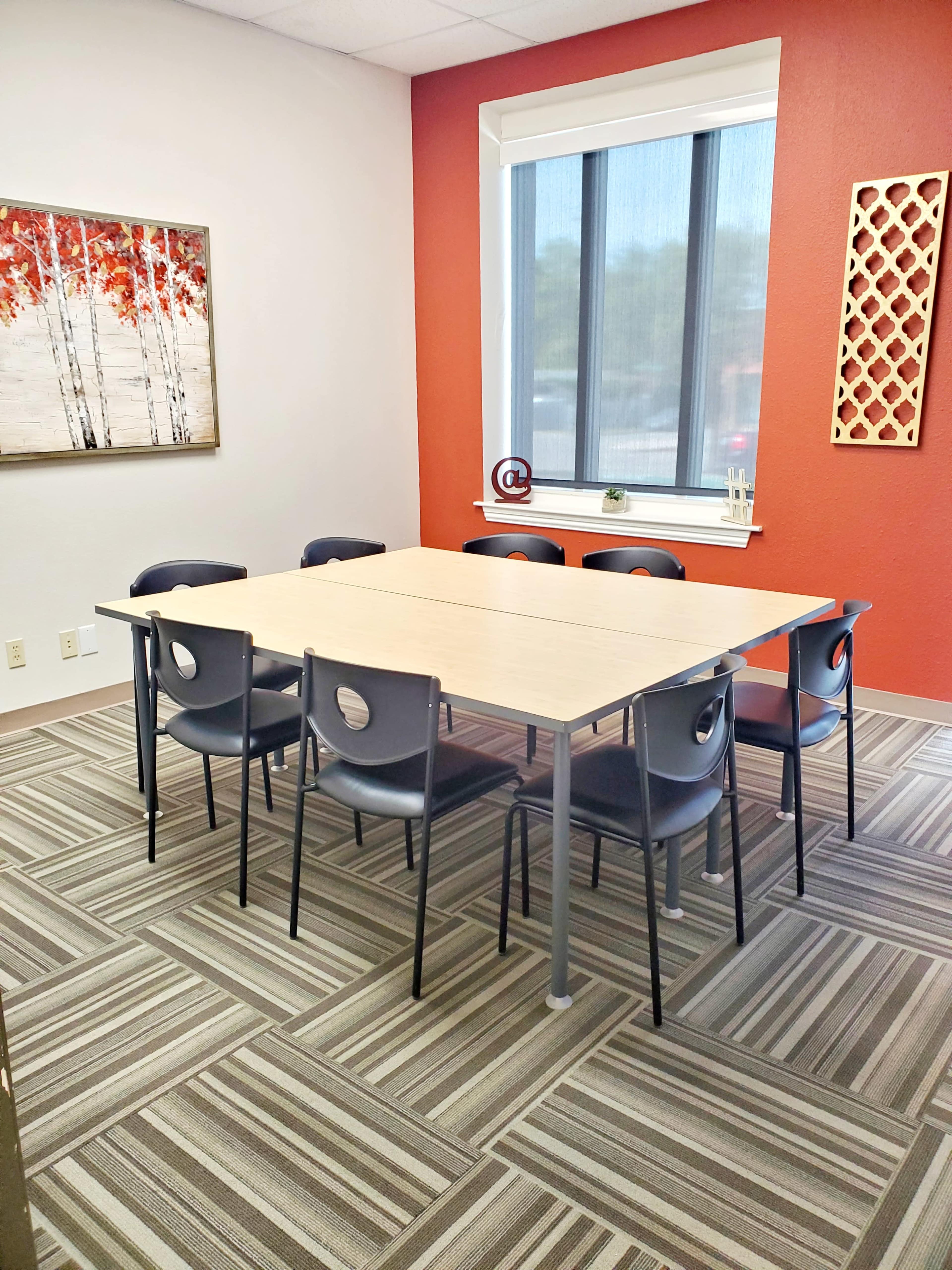 A modern meeting room features a rectangular table surrounded by eight chairs, with a window and artwork on the walls.