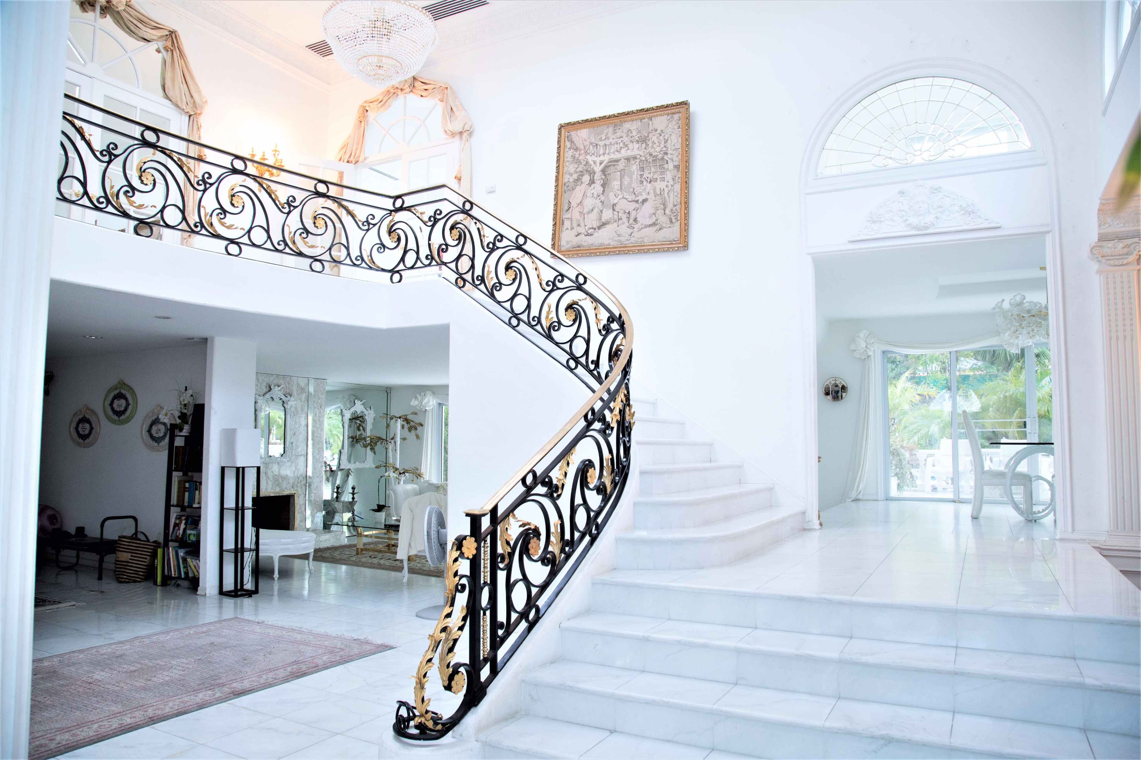 A spacious, elegant foyer with a sweeping staircase featuring ornate black and gold railings, leading to a bright, airy living area.