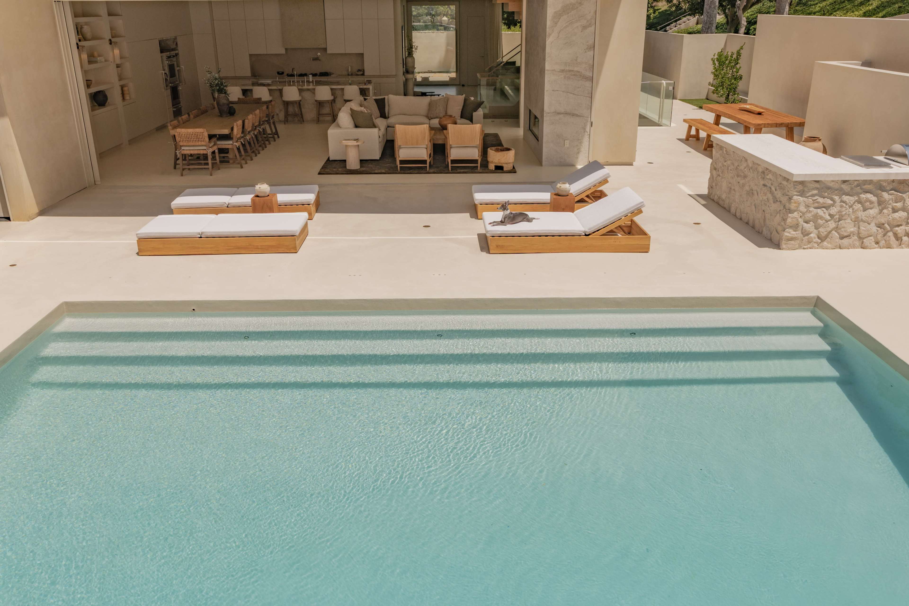 The image shows a modern outdoor space featuring a swimming pool with steps leading down to its clear water, lounge chairs along the edge, and an adjacent open-air kitchen area.