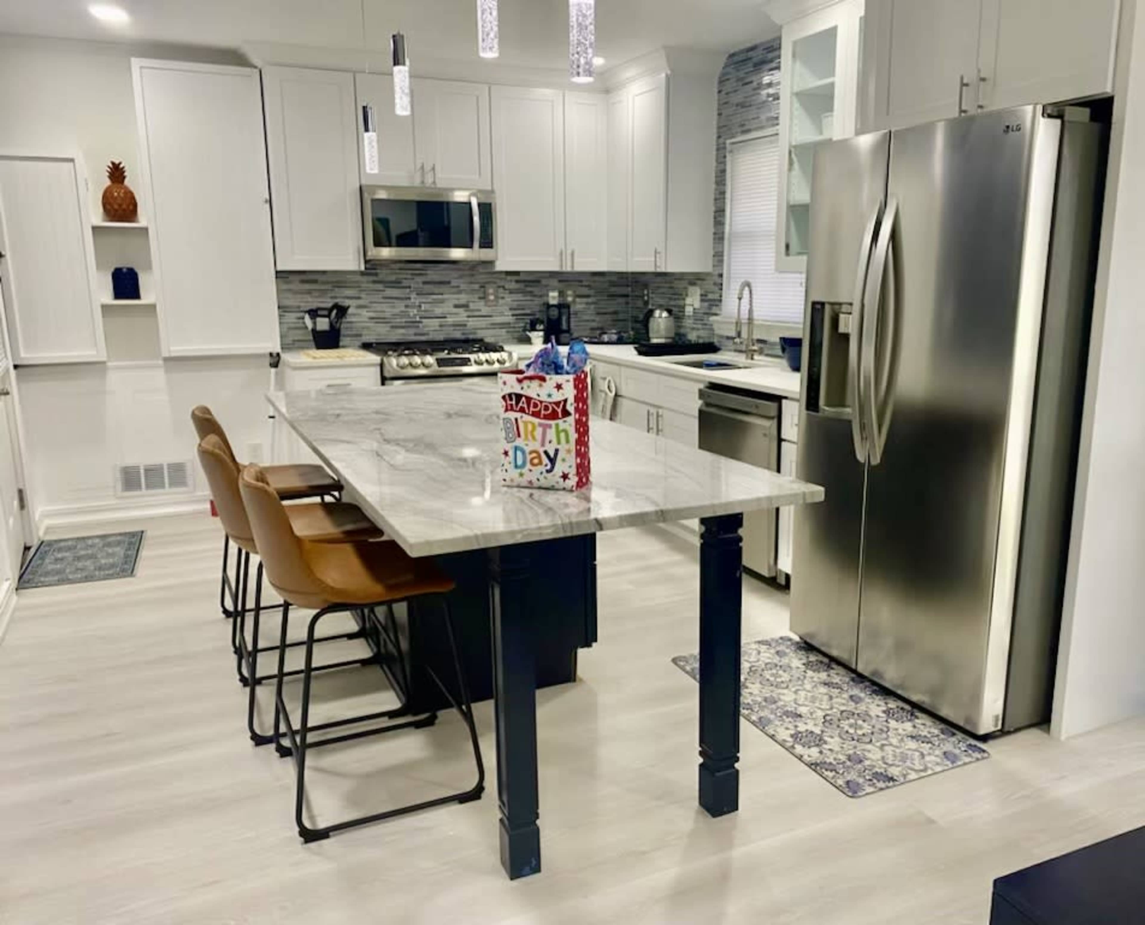A modern kitchen features white cabinetry, stainless steel appliances, and a marble island with four bar stools.